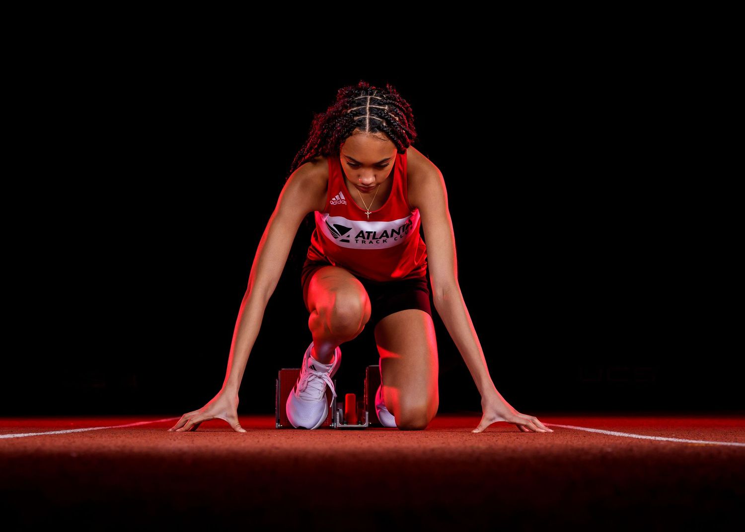 Athlete in red uniform crouches in starting position on a red track, ready for competition in dramatic lighting.