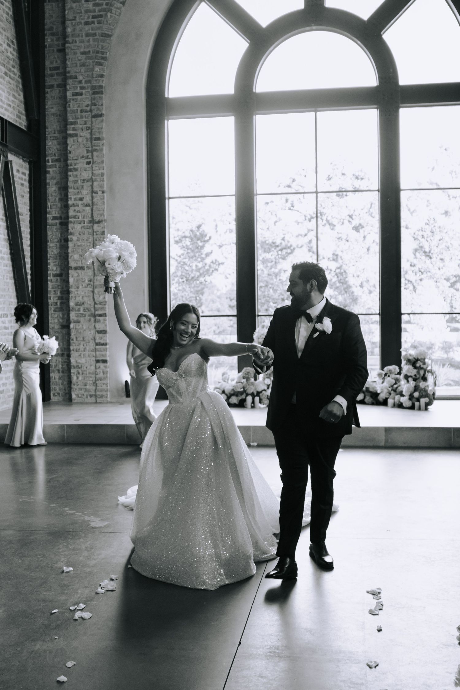 A wedding couple dances together in an elegant brick venue with arched windows and natural light streaming in.