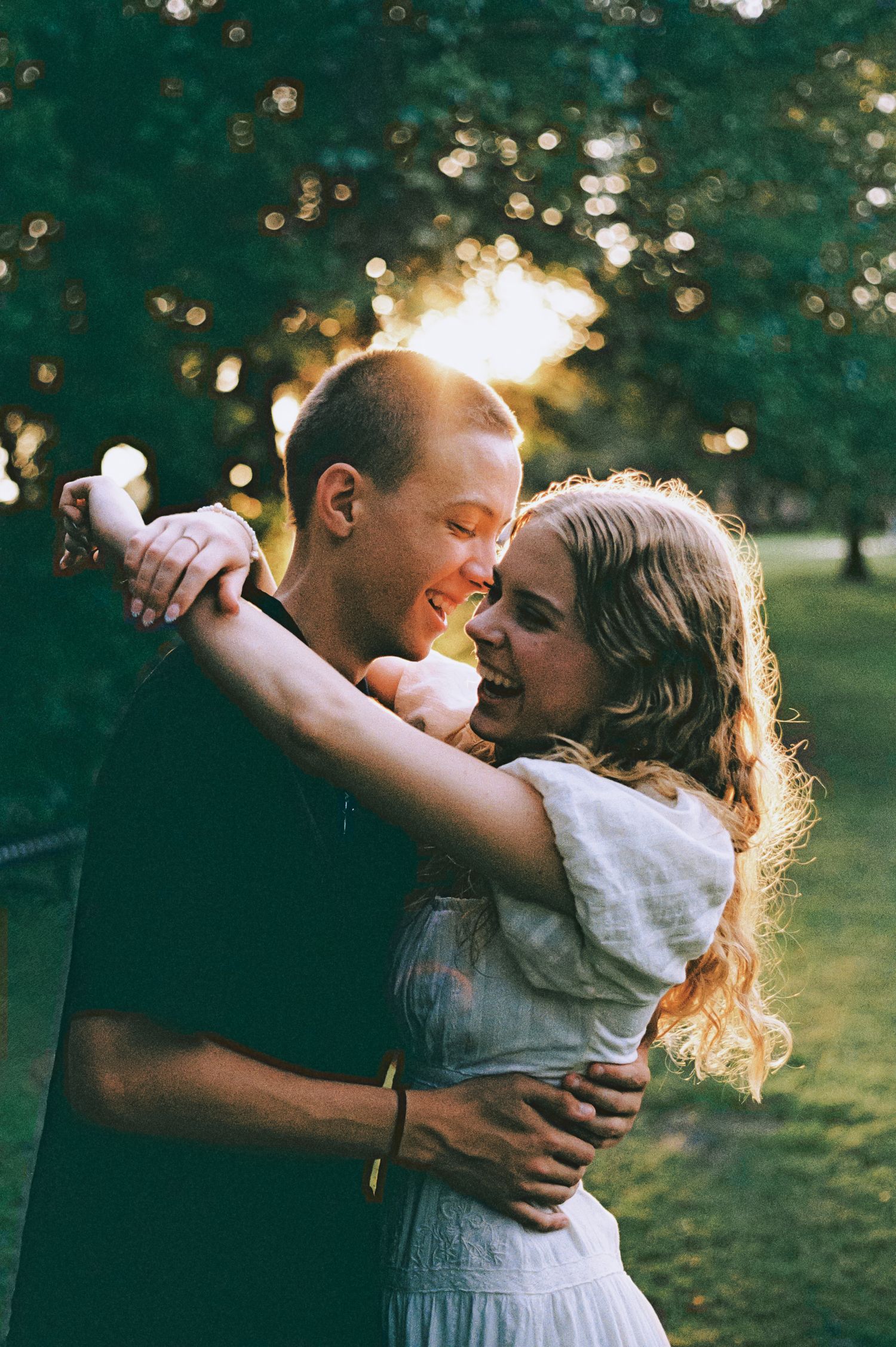 A joyful couple embraces and laughs together in a sunlit park with bokeh lights in the background.