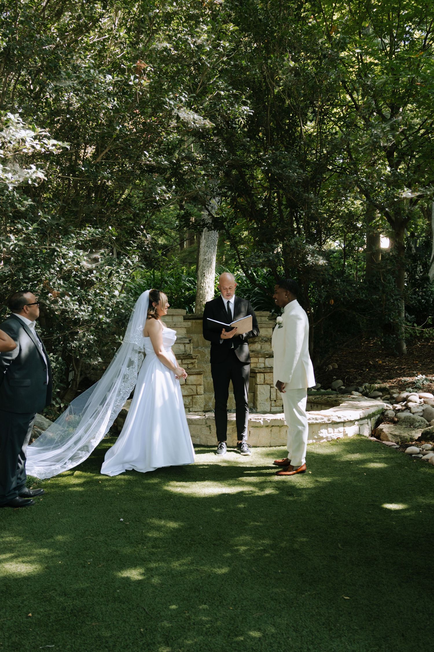 A wedding ceremony takes place in a lush garden setting with an officiant and couple standing beneath trees.