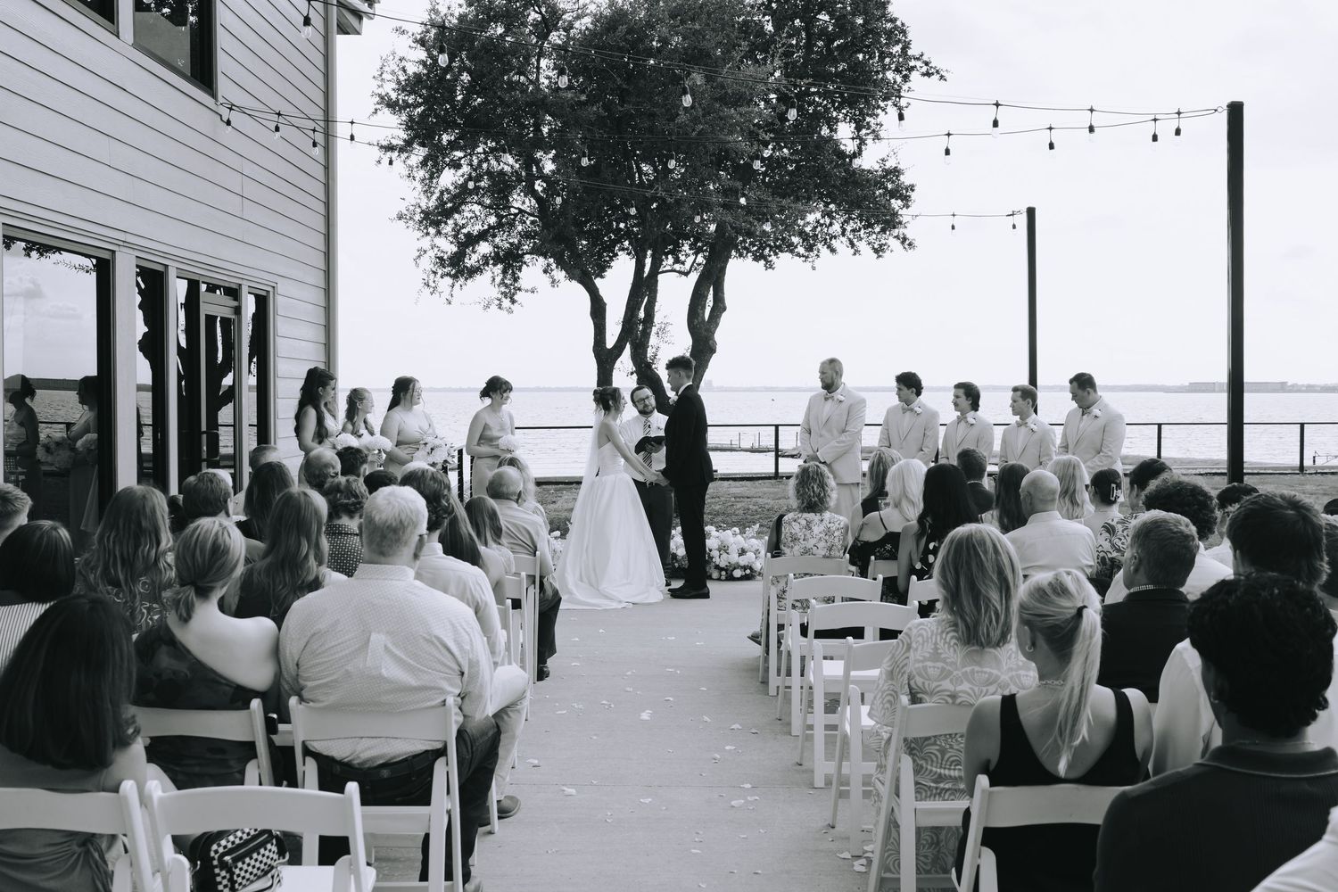 A series of black and white photos showing an outdoor wedding ceremony with guests seated in white chairs overlooking the water.
