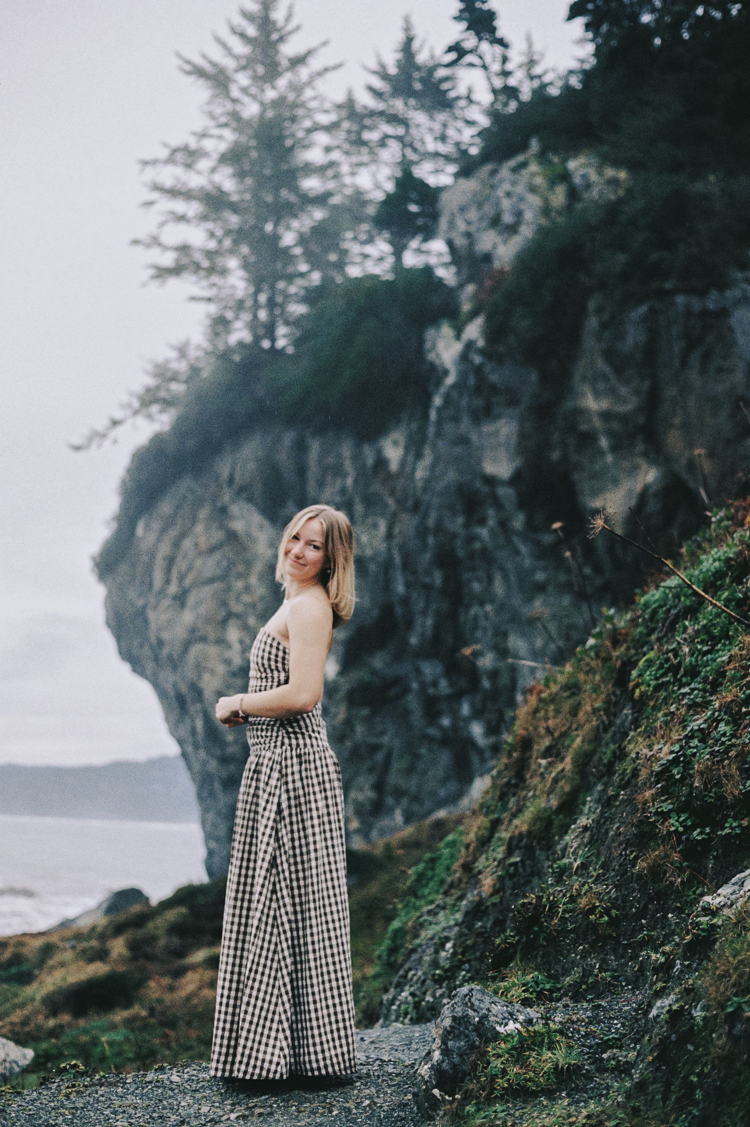 A person in a gingham dress stands contemplatively on a misty Pacific Northwest beach with dramatic cliffs and evergreens.