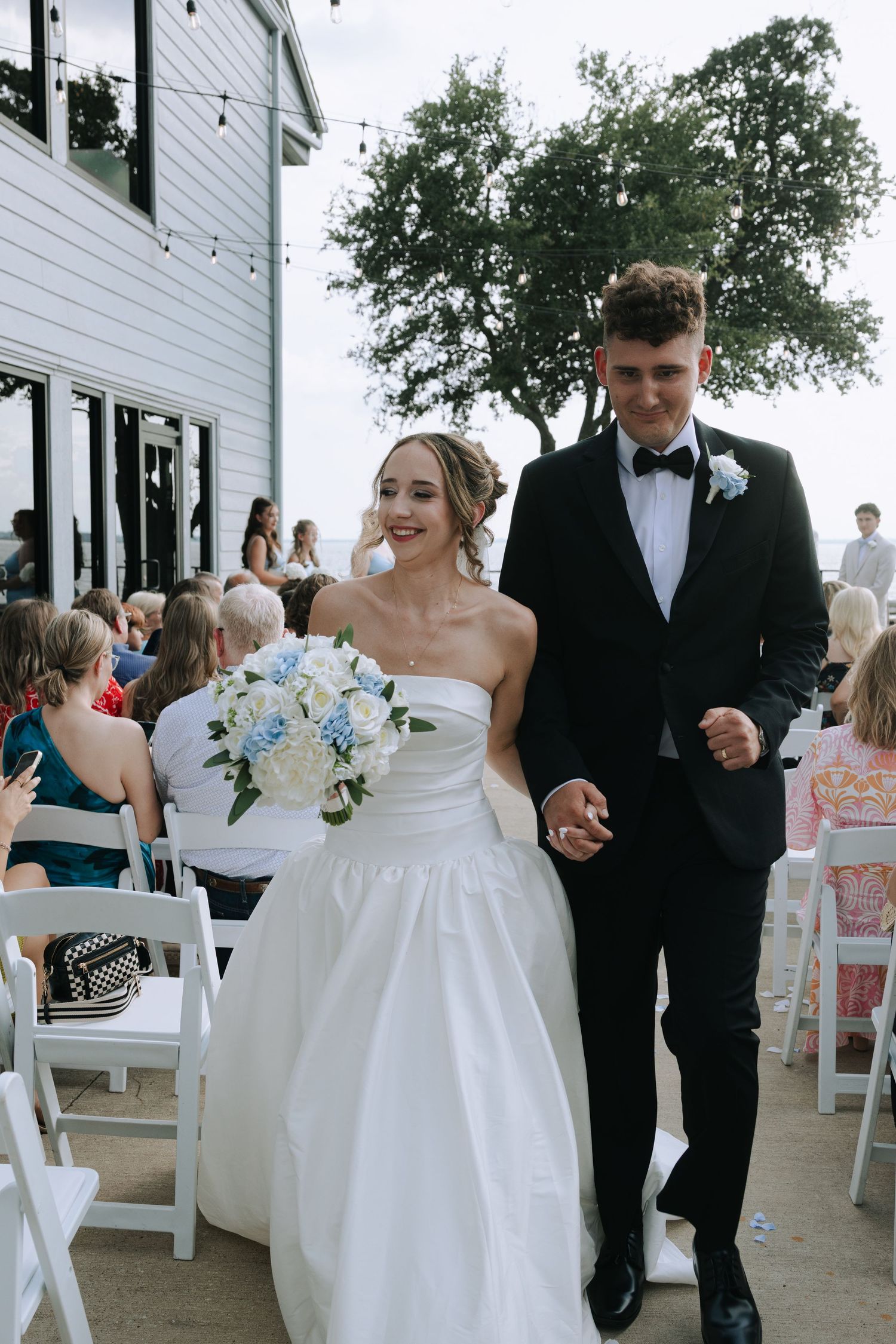 Wedding party members walk down an outdoor aisle beside a white building with guests seated on white chairs.