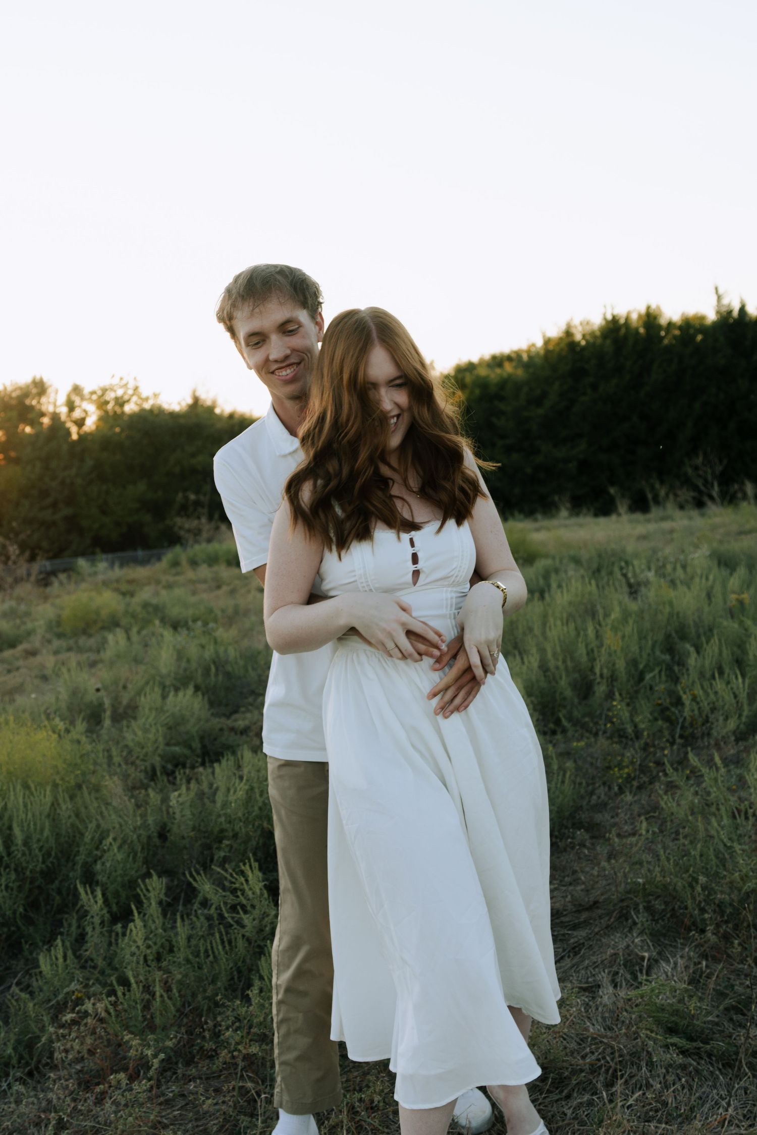 A couple in white and khaki clothing embraces lovingly in a grassy field at sunset.