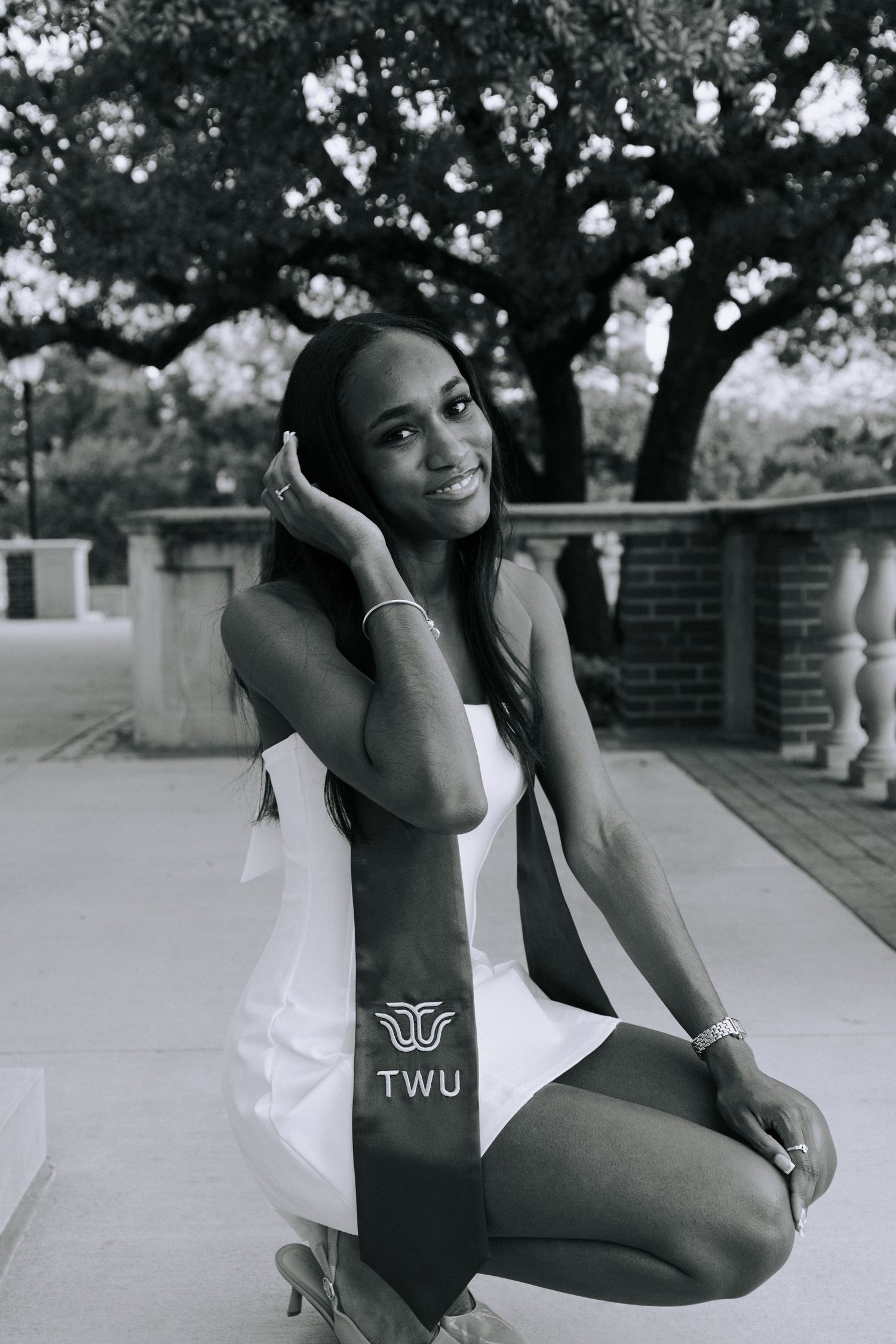 Black and white portrait of a person in TWU athletic wear posing outdoors near a tree.