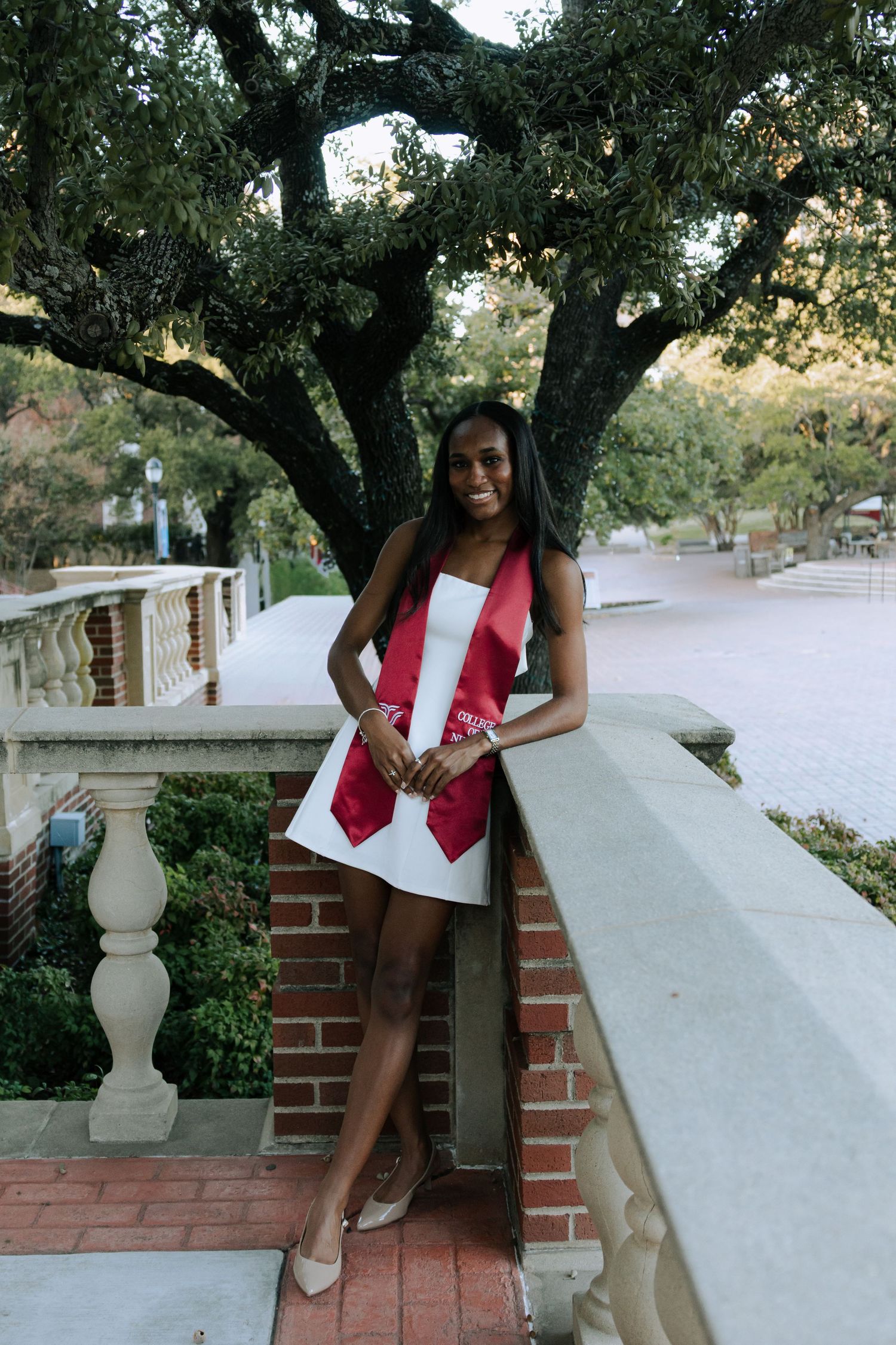 A person in a red and white outfit poses on a brick wall under trees on a sunny day.