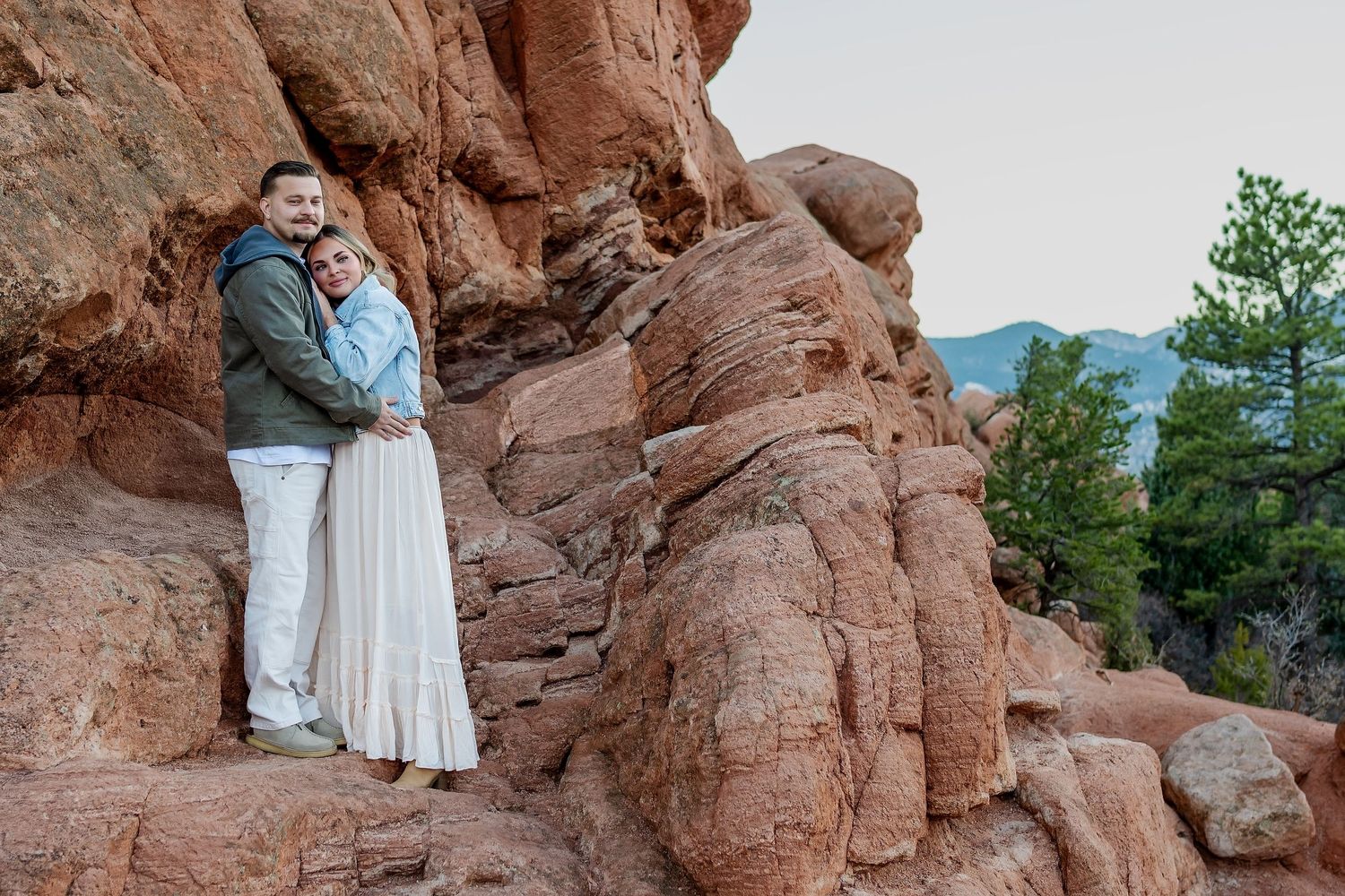 Couple standing together on red rocks with pine trees in background.