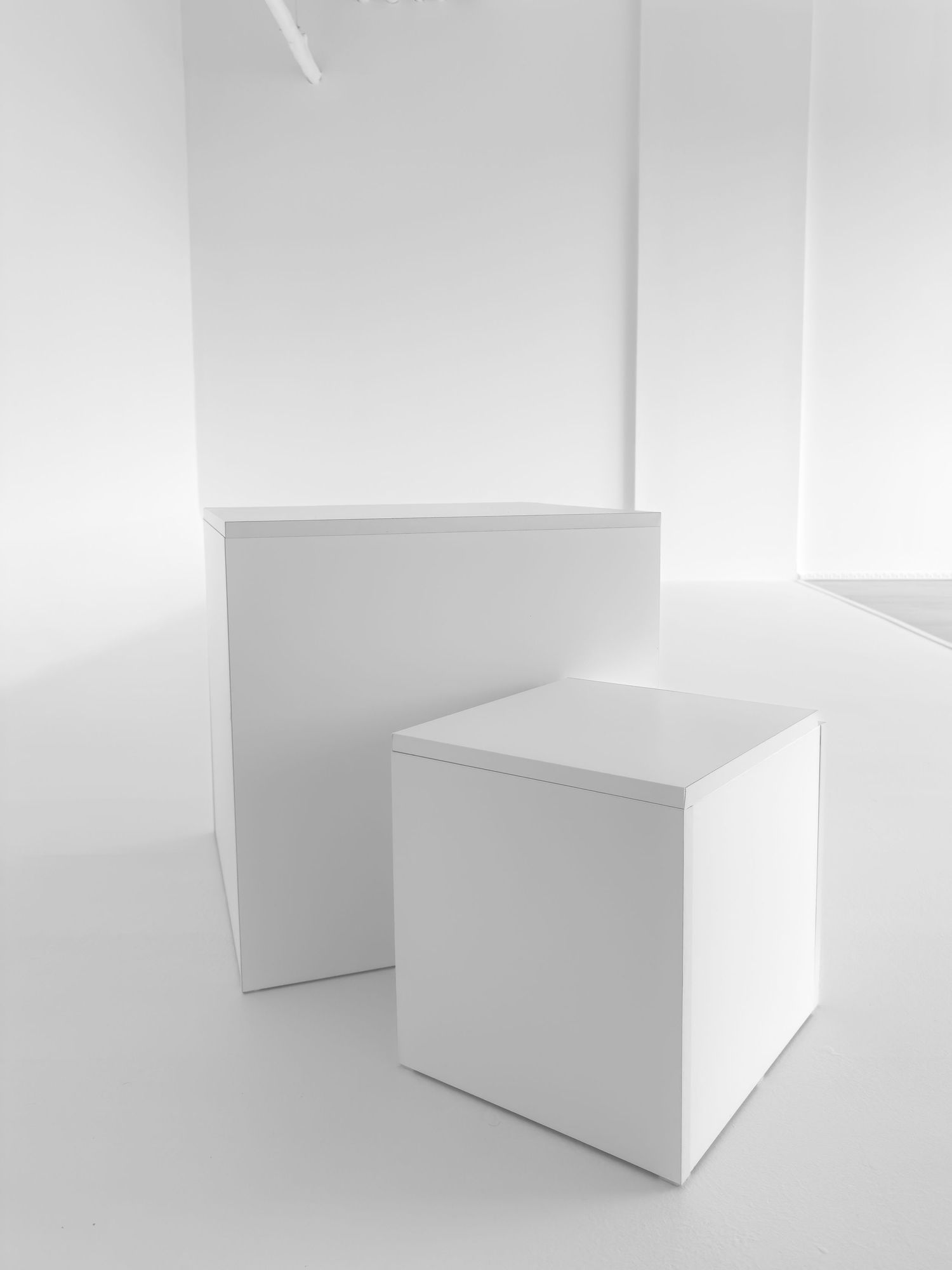 Two white minimalist cube pedestals of different heights displayed in a bright white gallery space.