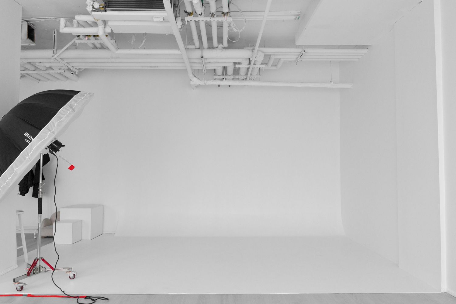 A bright white photography studio with seamless backdrop, professional lighting umbrella, and exposed ceiling pipes.