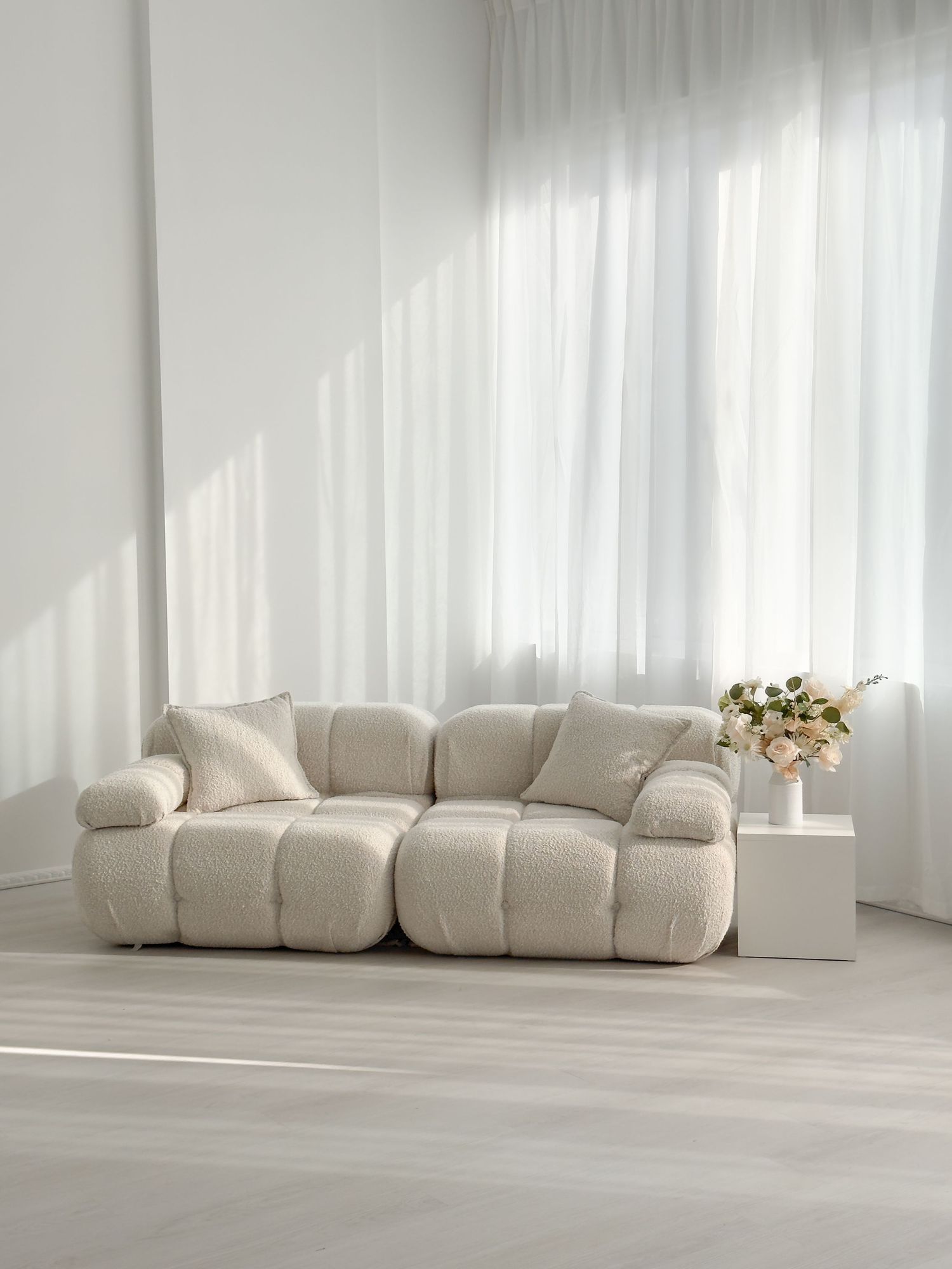 Minimalist living room with a cream bouclé modular sofa, sheer white curtains, and a small side table with flowers.