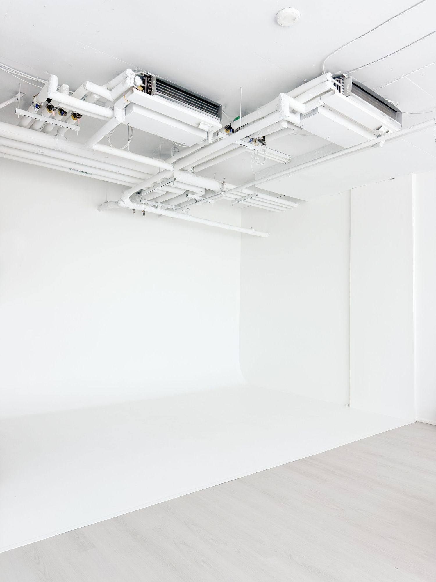 Minimalist white room with exposed ceiling ductwork, pipes, and track lighting in a clean modern interior space.