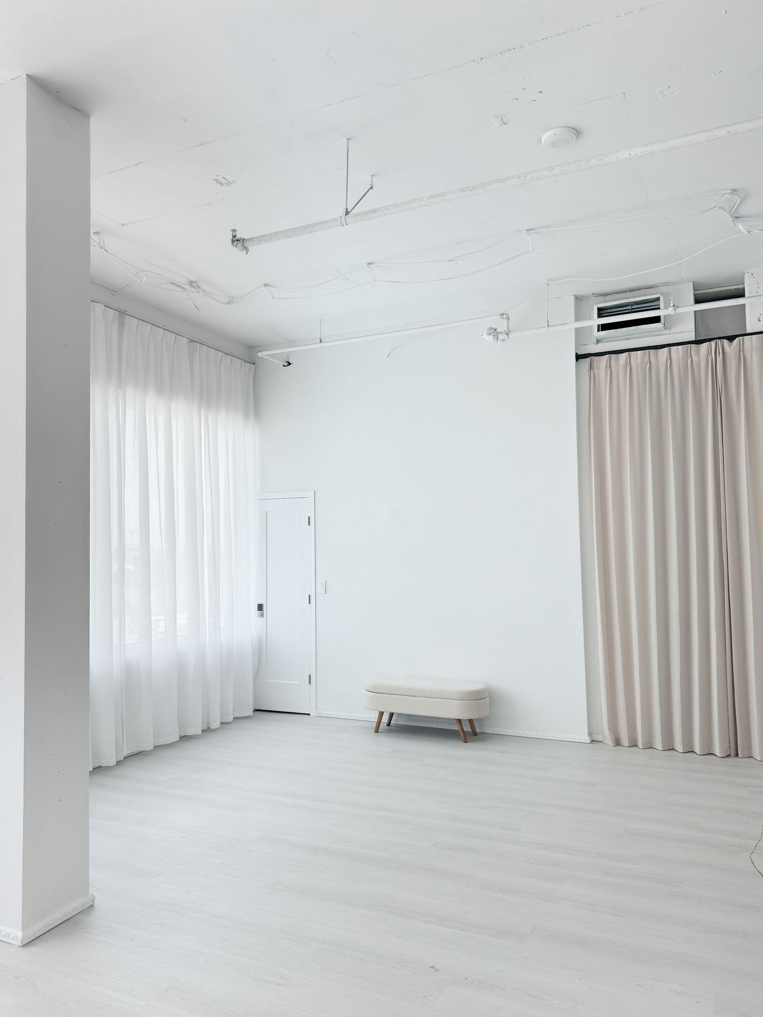 Minimalist white studio space with sheer curtains, beige drapes, light wood floors, and a small upholstered bench.