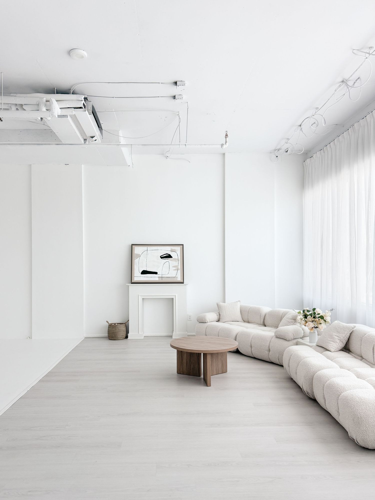 Minimalist white living room with modular cream sofa, round wood coffee table, white fireplace, and sheer curtains.