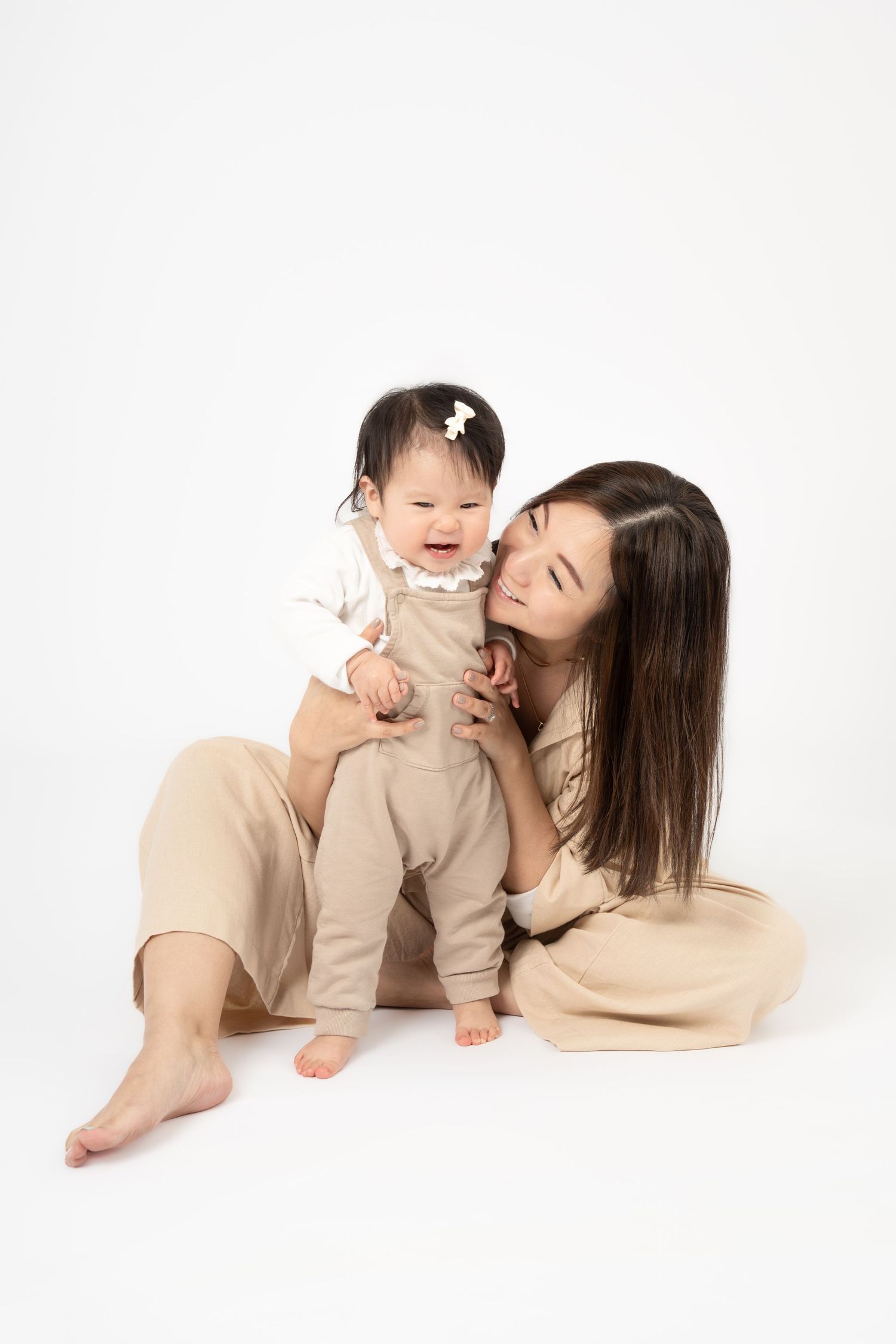 A young Asian mother sitting on white background, smiling while holding her laughing baby girl in beige overalls.