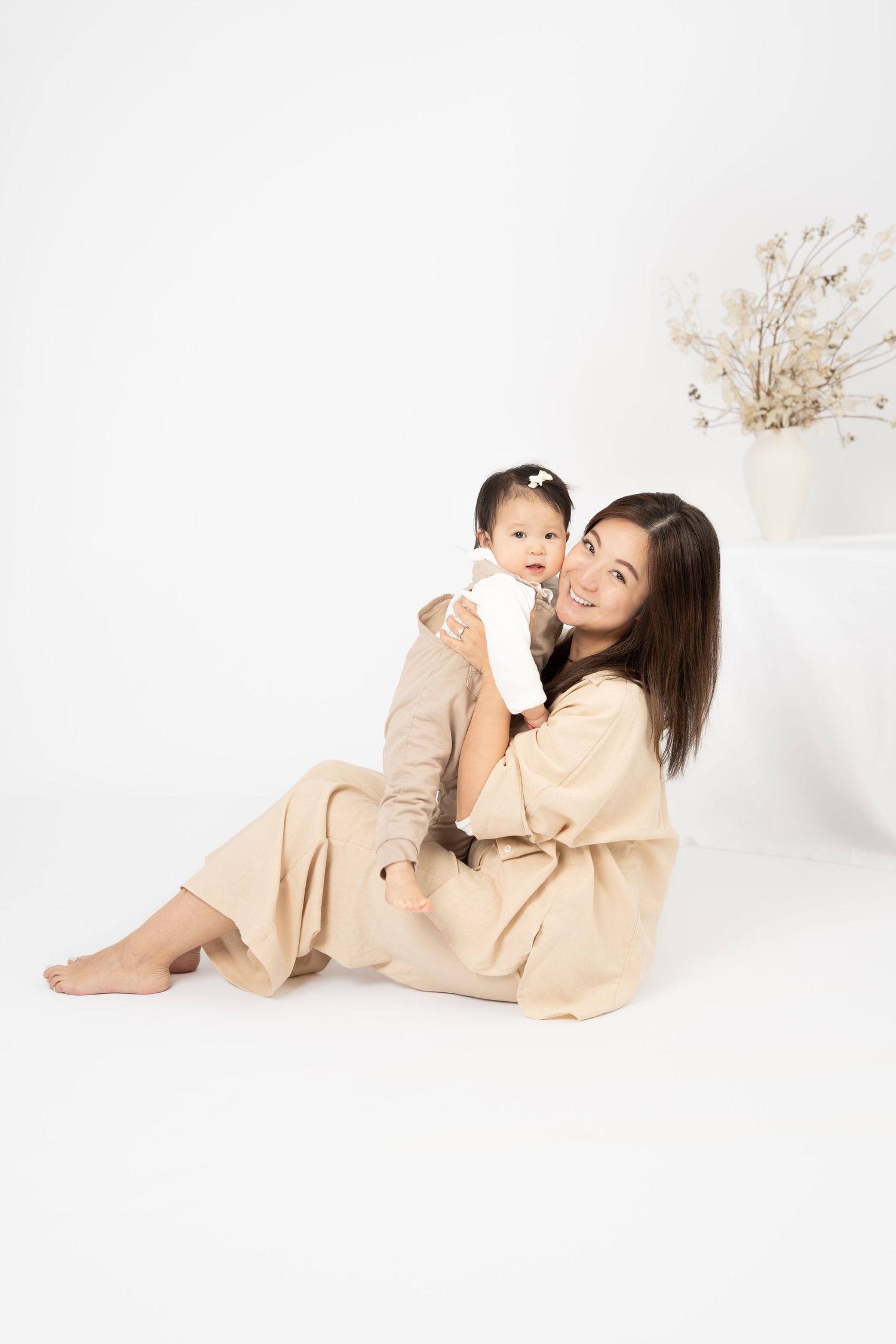 A smiling Asian mother in beige outfit sits on white floor holding her baby girl in a bright studio setting.