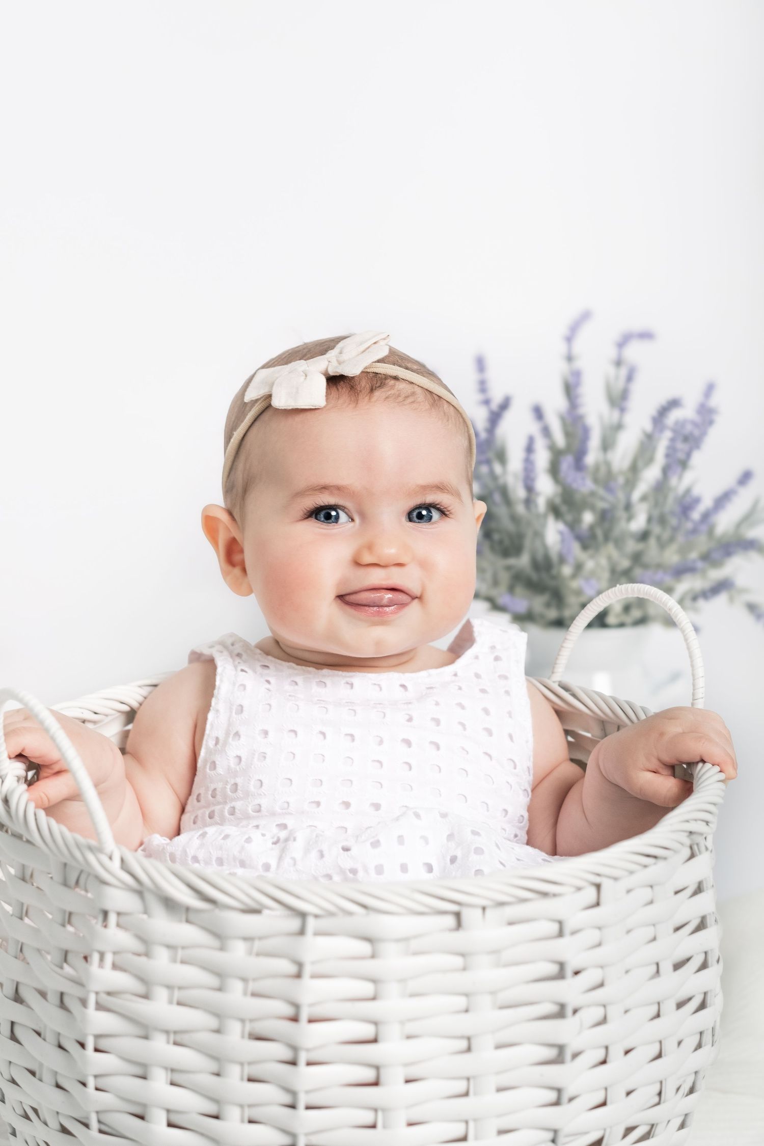 Baby wearing white clothing sits in a woven basket with lavender flowers in the background.