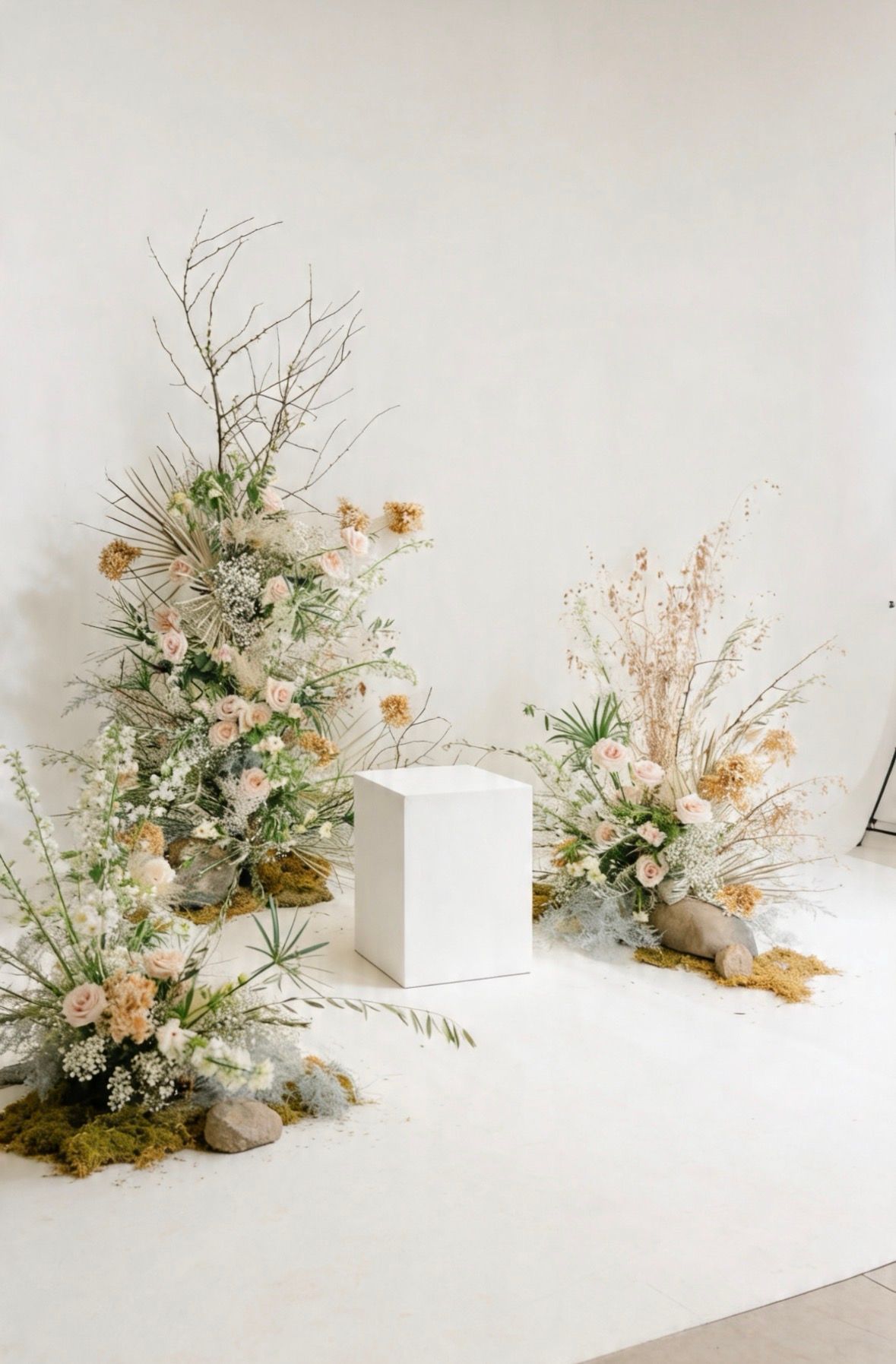 Elegant floral arrangements with dried botanicals, blush blooms, and greenery flanking a white pedestal on a neutral backdrop.