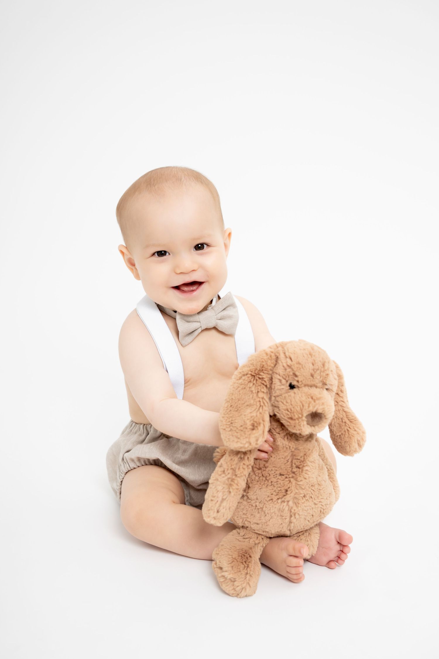 Baby in neutral outfit playing with soft plush puppy toy on white background.