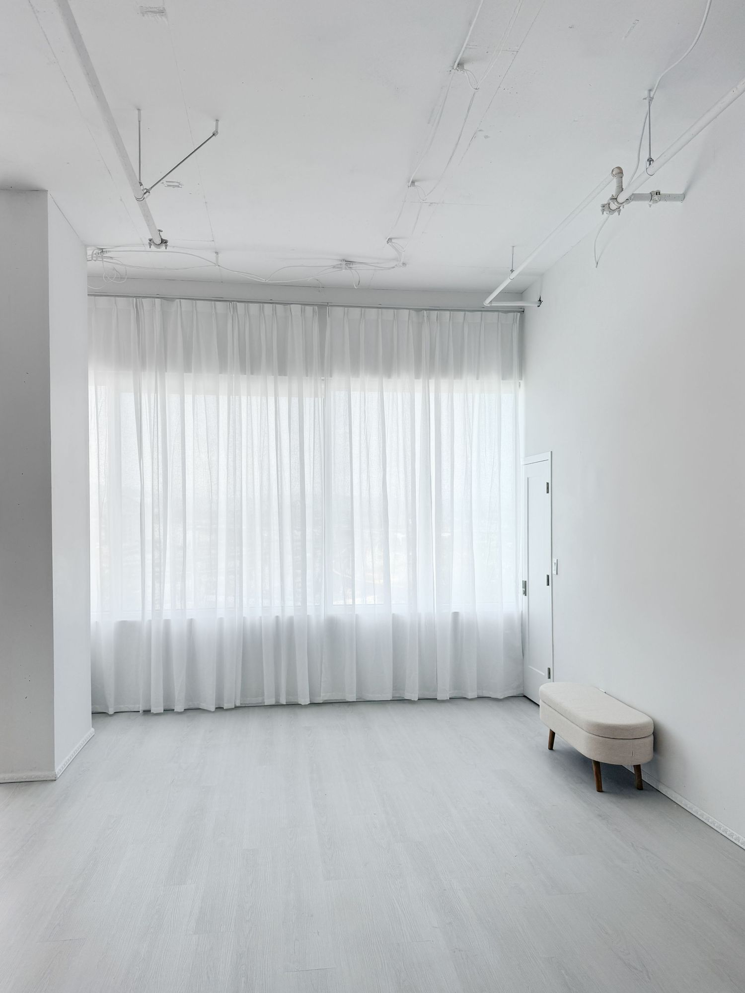 Minimalist all-white room with sheer curtains, light wood floors, and a small upholstered bench.