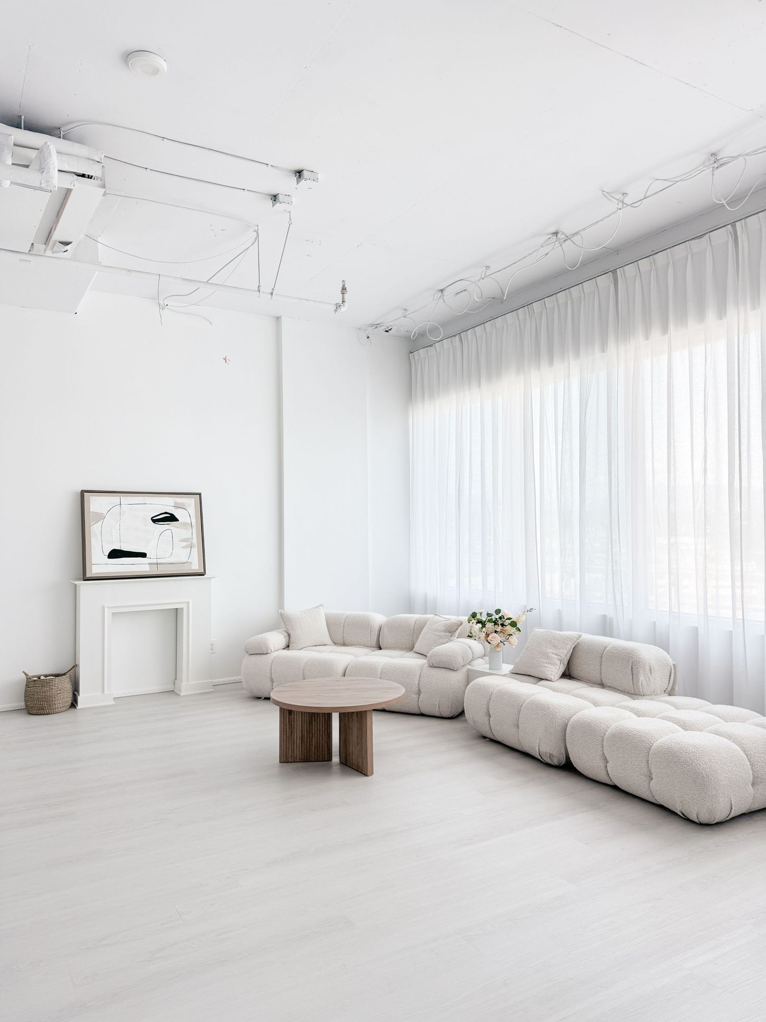 Minimalist white living room with a tufted cream sofa, round wood coffee table, sheer curtains, and abstract wall art.