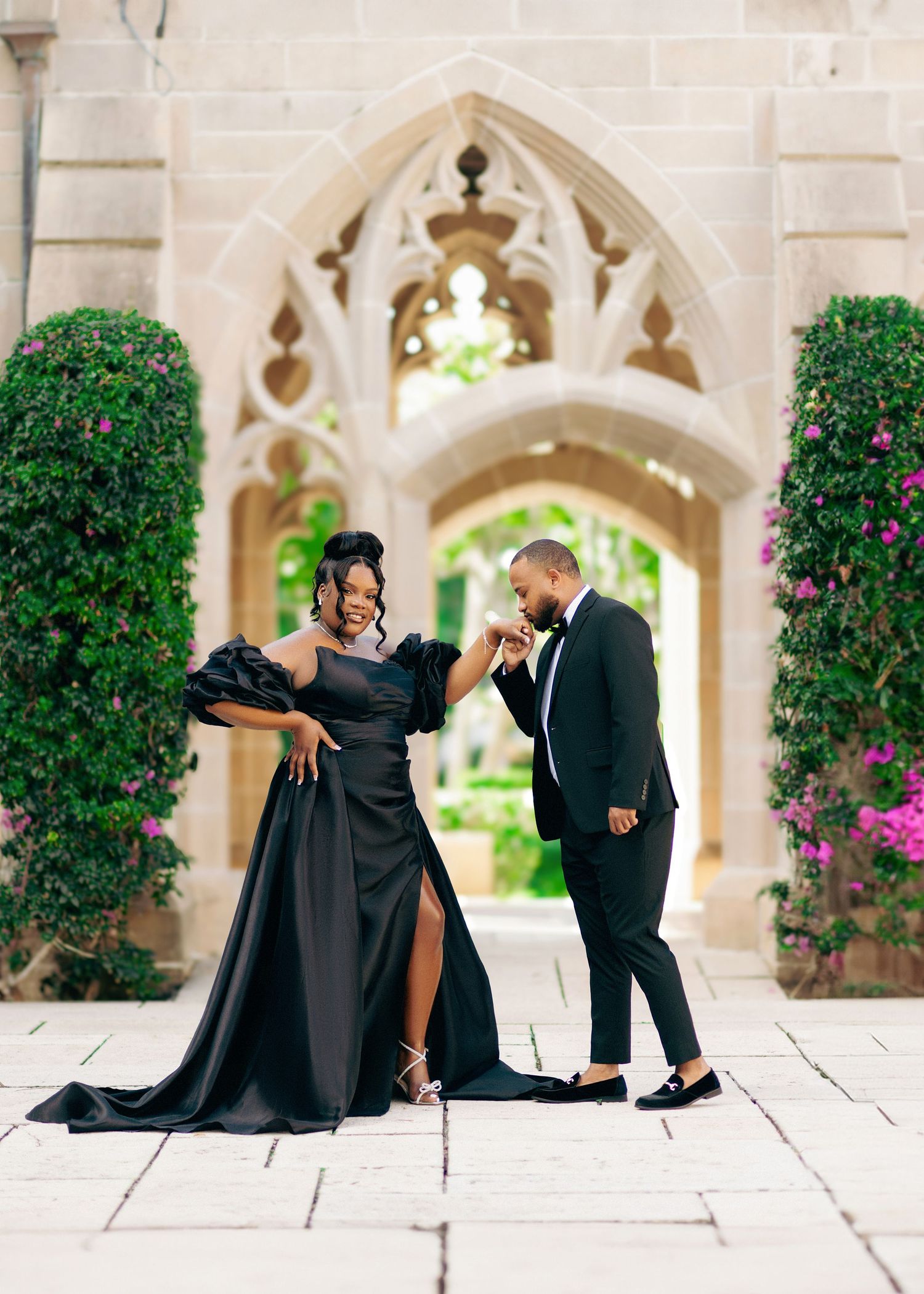Couple in elegant black formal wear pose at a gothic stone archway surrounded by lush greenery and pink flowers.