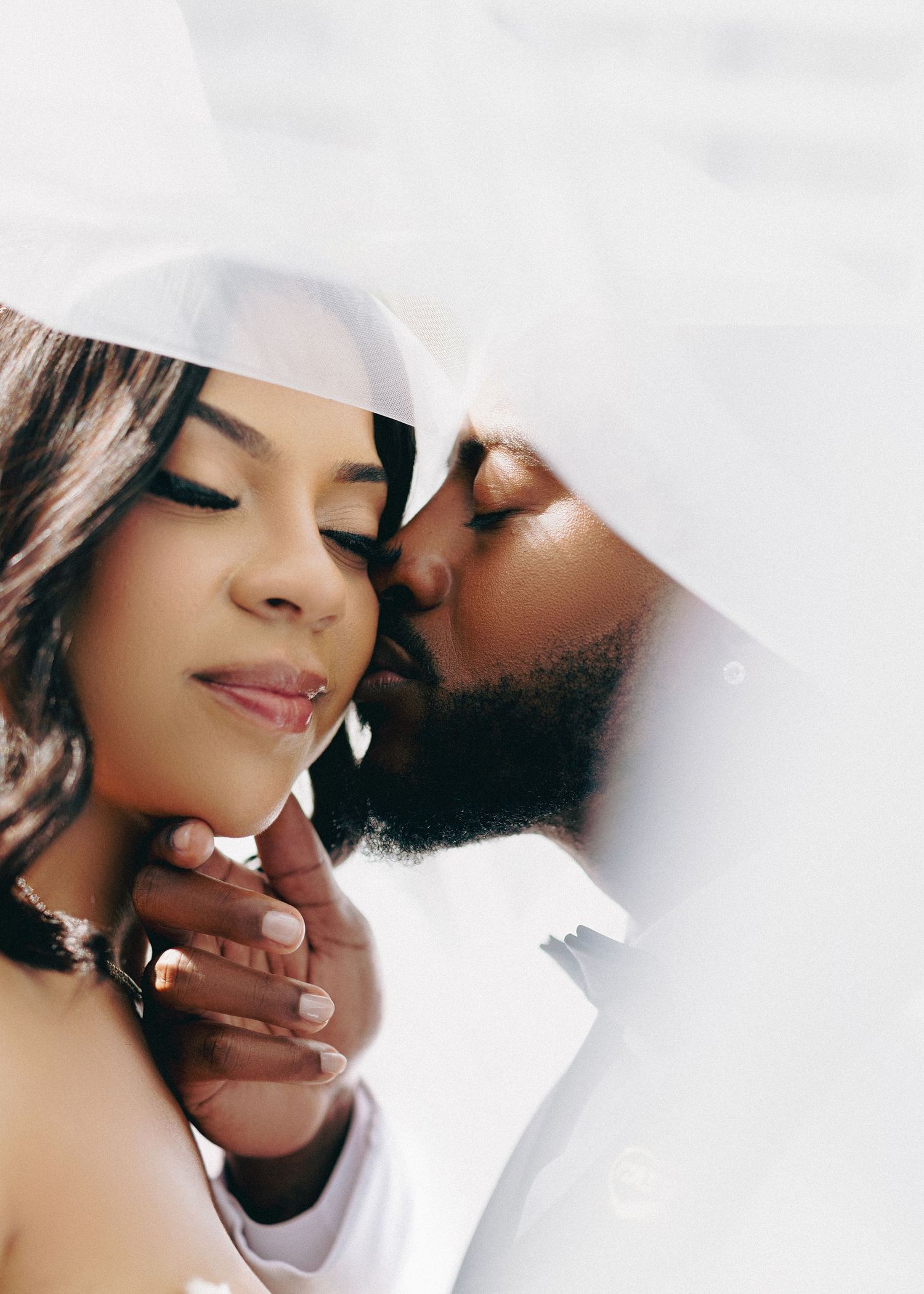 A romantic couple shares a tender kiss under a white veil during their wedding day photoshoot.