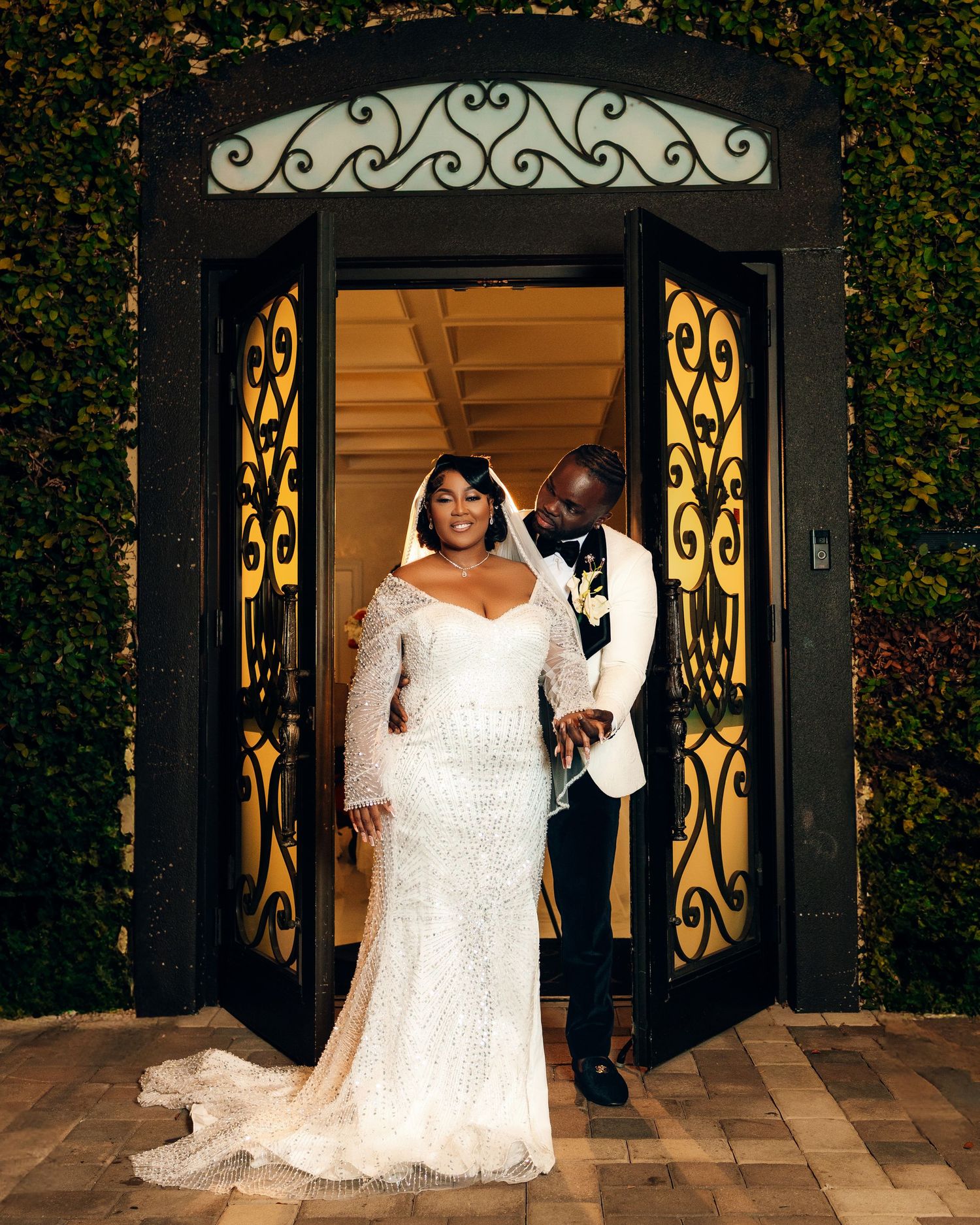 Elegant wedding couple poses in front of ornate iron gates illuminated by warm evening lighting.