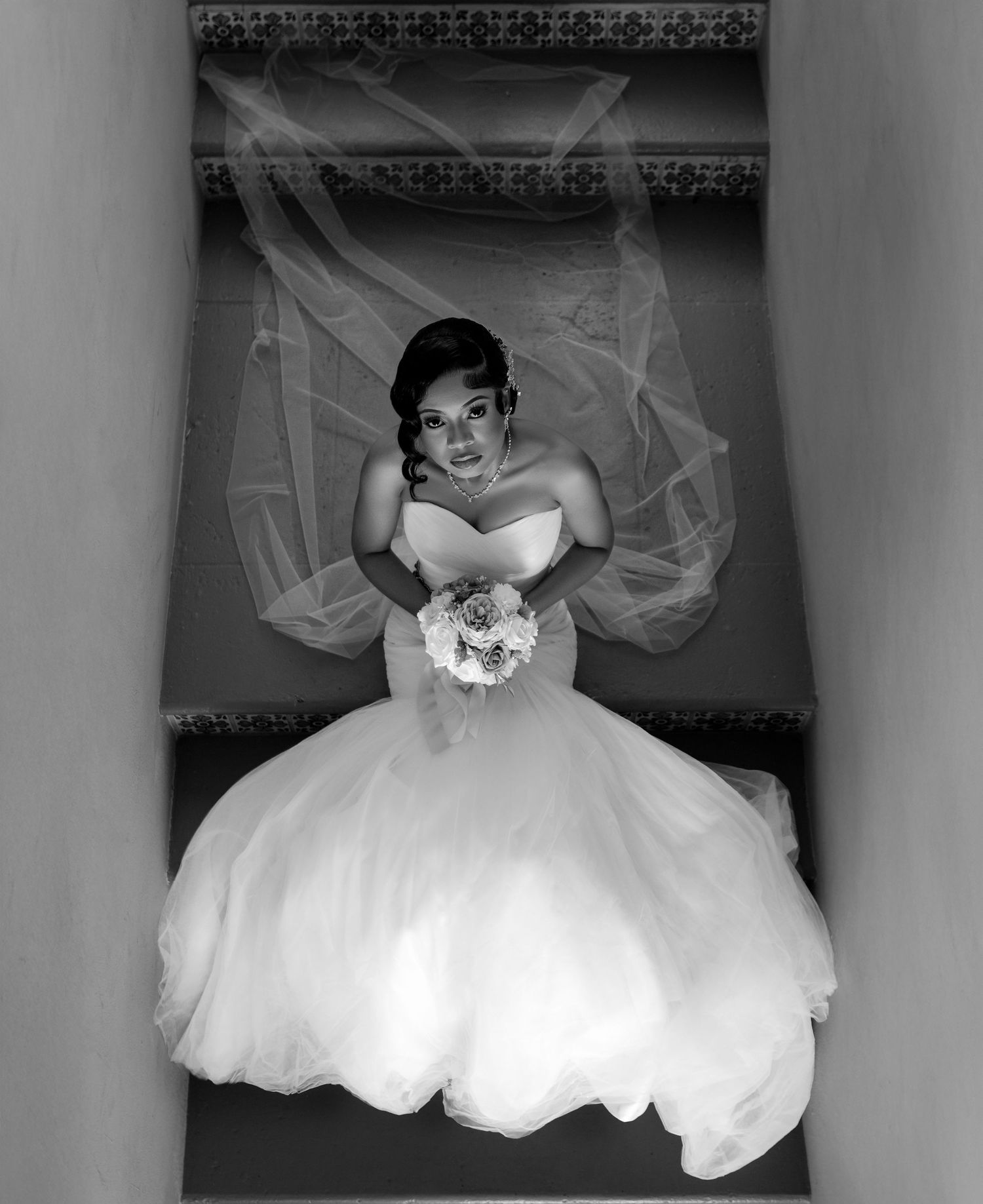 A bride in a voluminous white ball gown poses elegantly on a staircase in black and white photography.