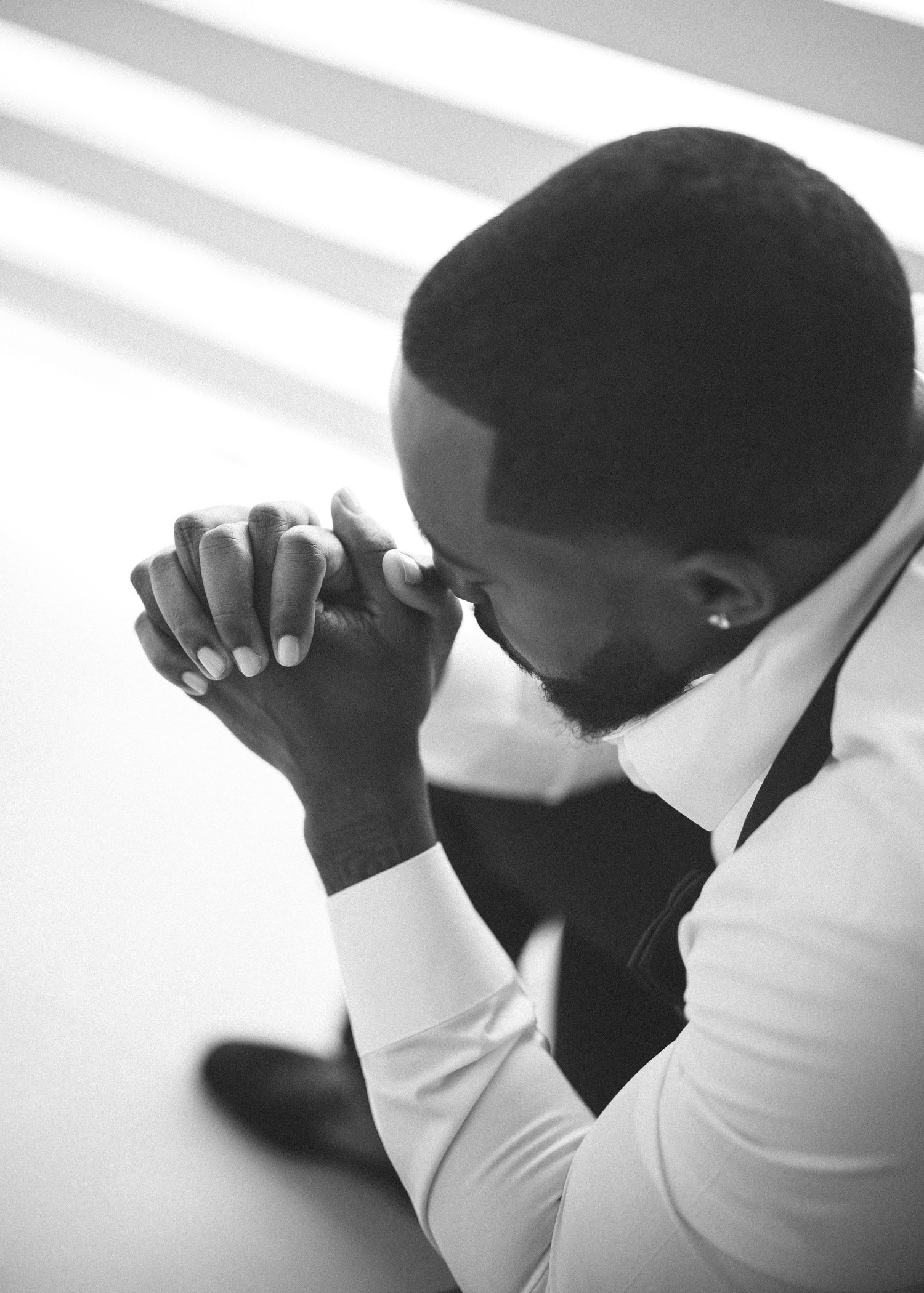 Groom in white shirt bows head in prayer with hands clasped before wedding ceremony.