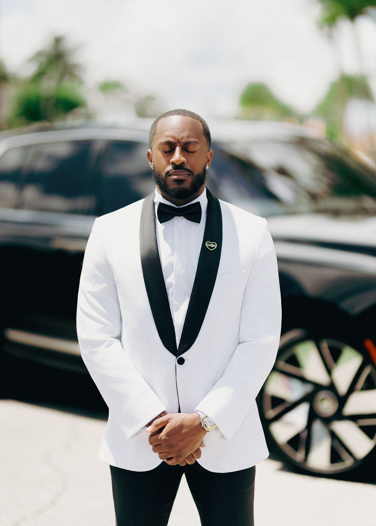 Groom in white tuxedo with black bow tie standing outdoors near a luxury black vehicle.