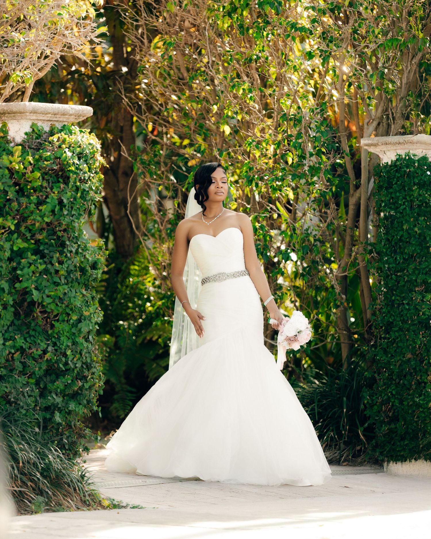 Elegant white wedding gown photographed in a garden setting with lush greenery and sunlit pathway.