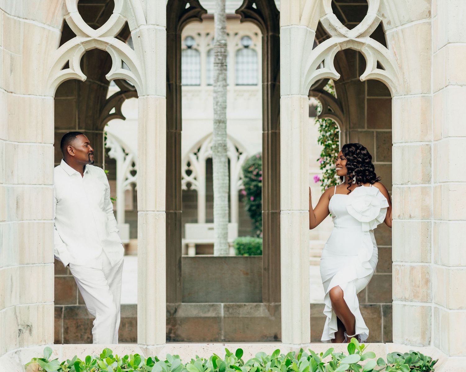 Couple in white attire embraces in front of ornate stone colonnade.