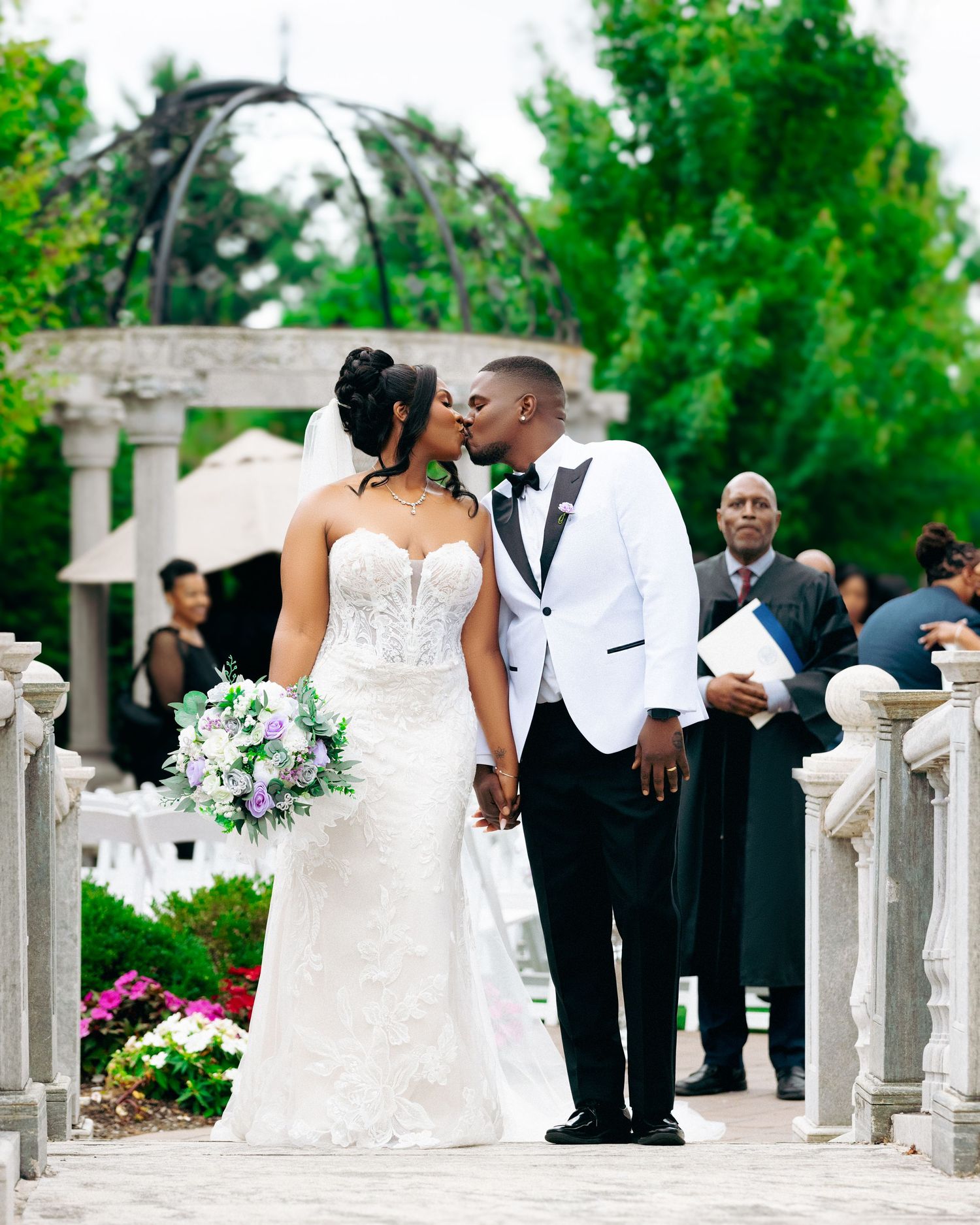 Wedding couple posing together along an ornate white stone balustrade in a garden setting.