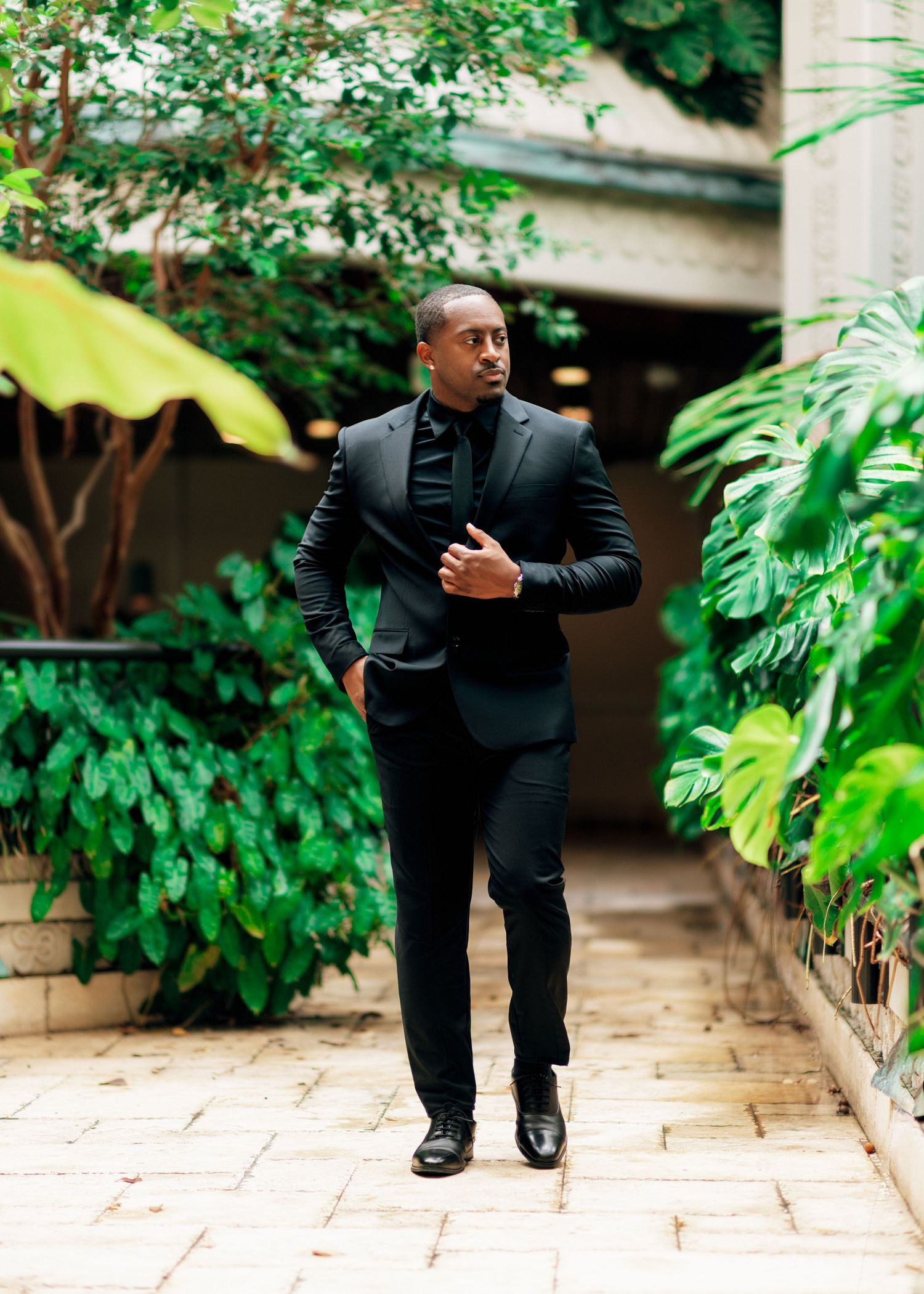 Person in black suit walking along garden pathway with lush greenery.