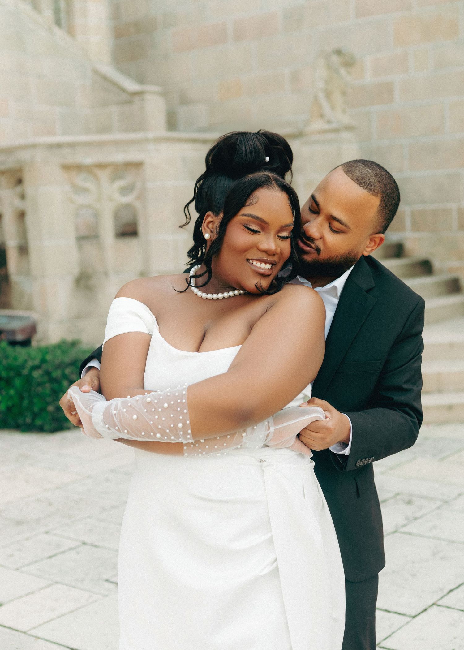 A tender moment between newlyweds embracing and smiling against a stone building backdrop.