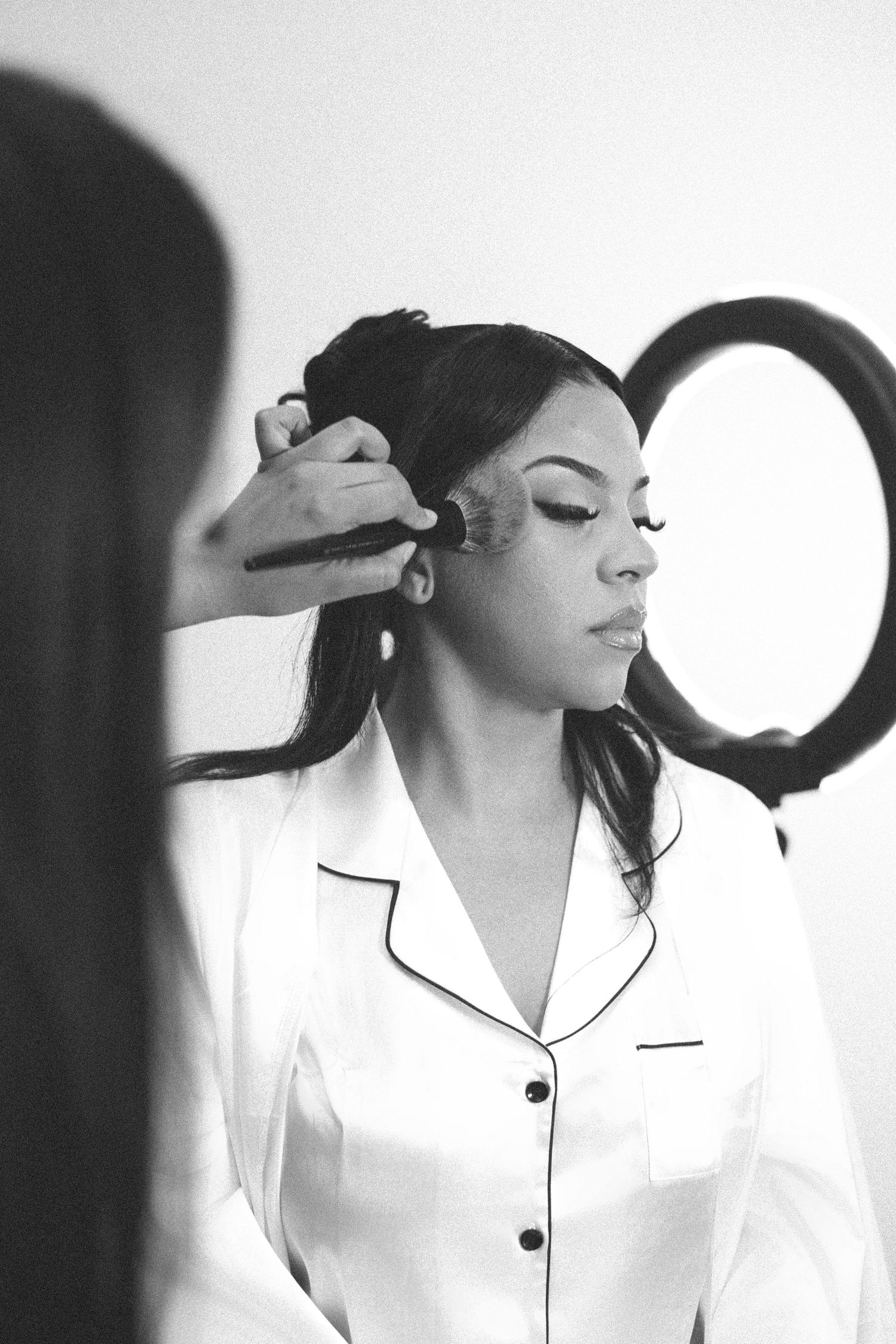 Black and white photo of a woman in silk pajamas having makeup applied by a makeup artist near a ring light.