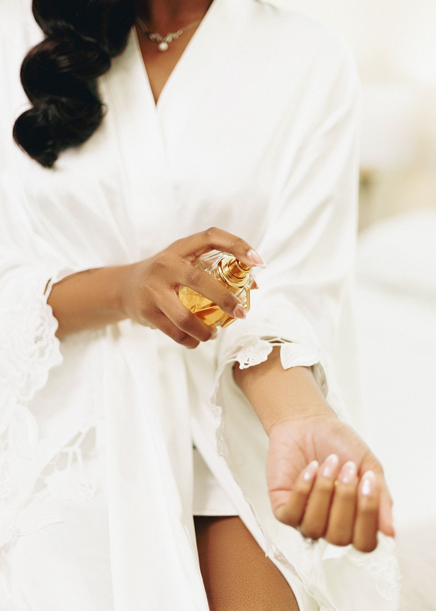 Close-up of hands wearing white satin robes applying perfume and jewelry while getting ready.