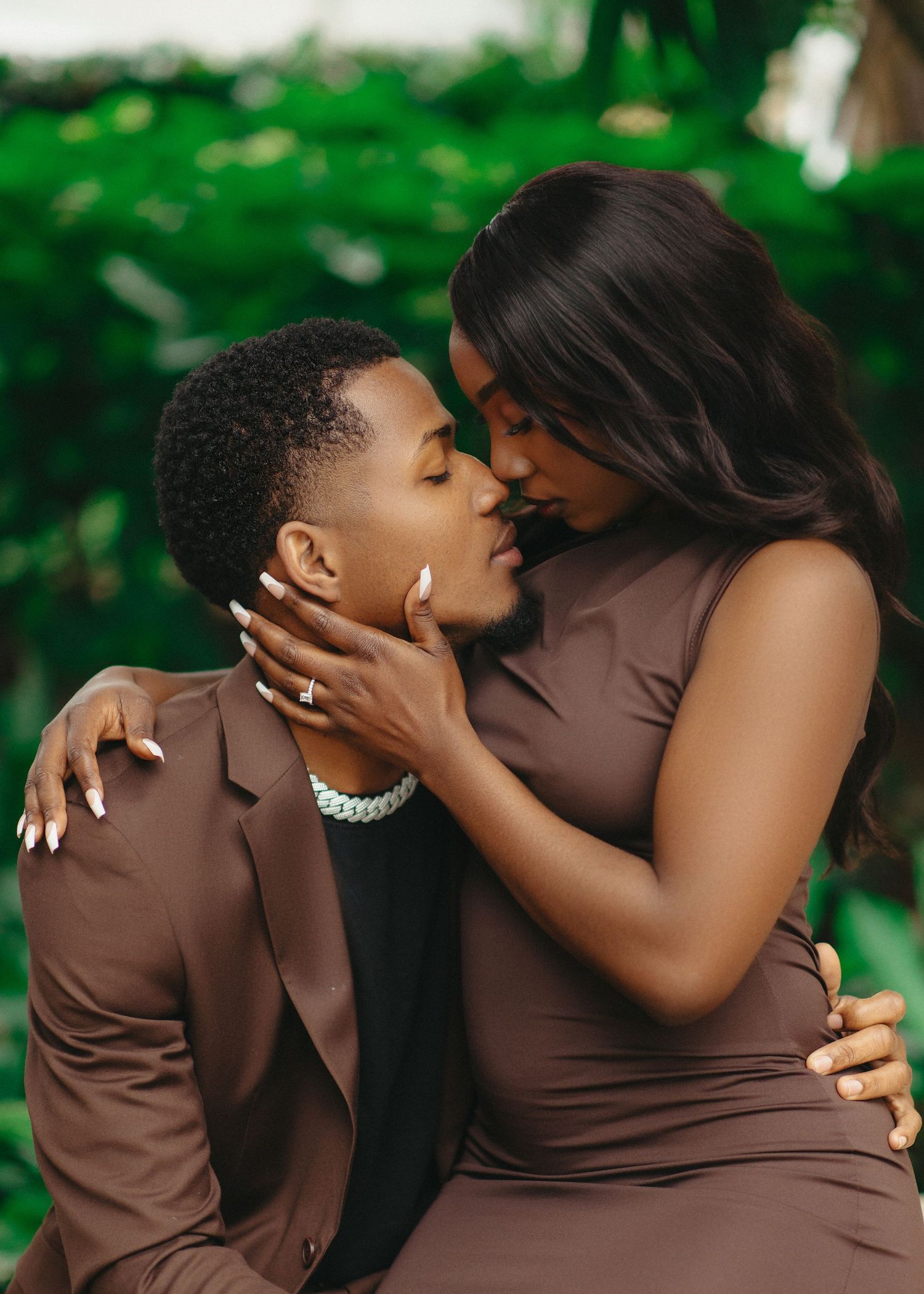 A couple in brown and black attire share an intimate moment in a lush green outdoor setting.