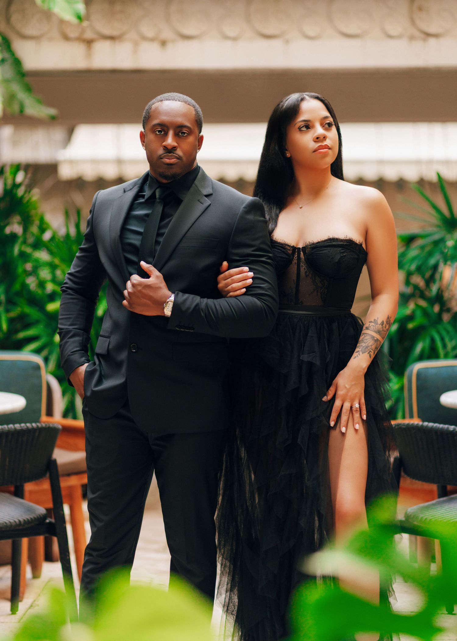 Couple in formal black attire posing together in tropical garden setting.