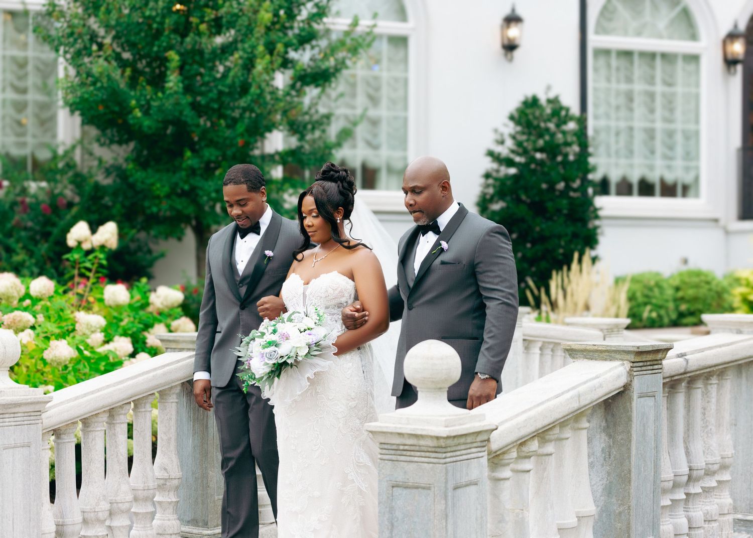 Wedding party gathered along a decorative white stone railing with lush greenery in the background.