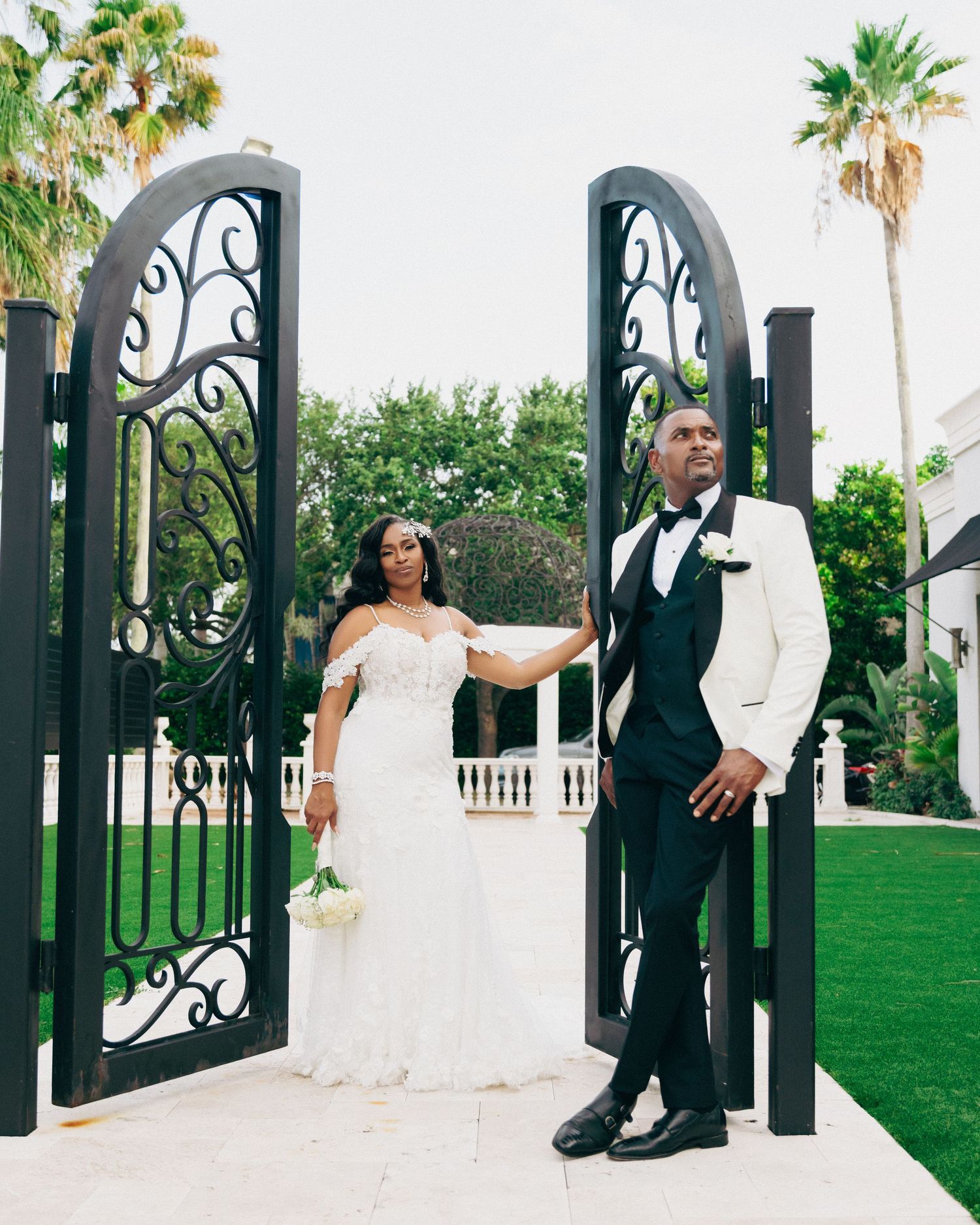 Wedding couple poses by ornate black iron gates with palm trees in background.