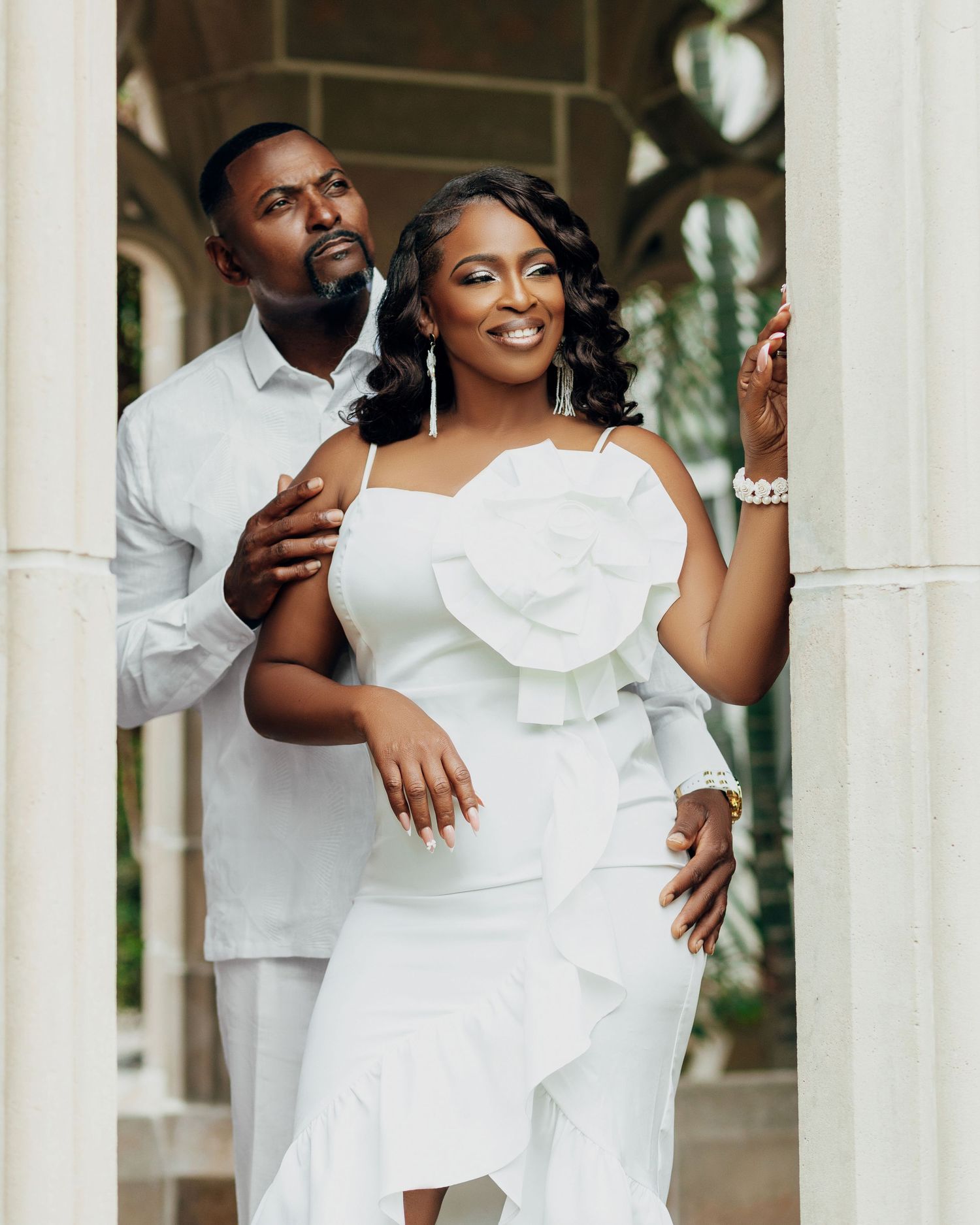 Couple in matching white outfits poses between classical stone pillars.