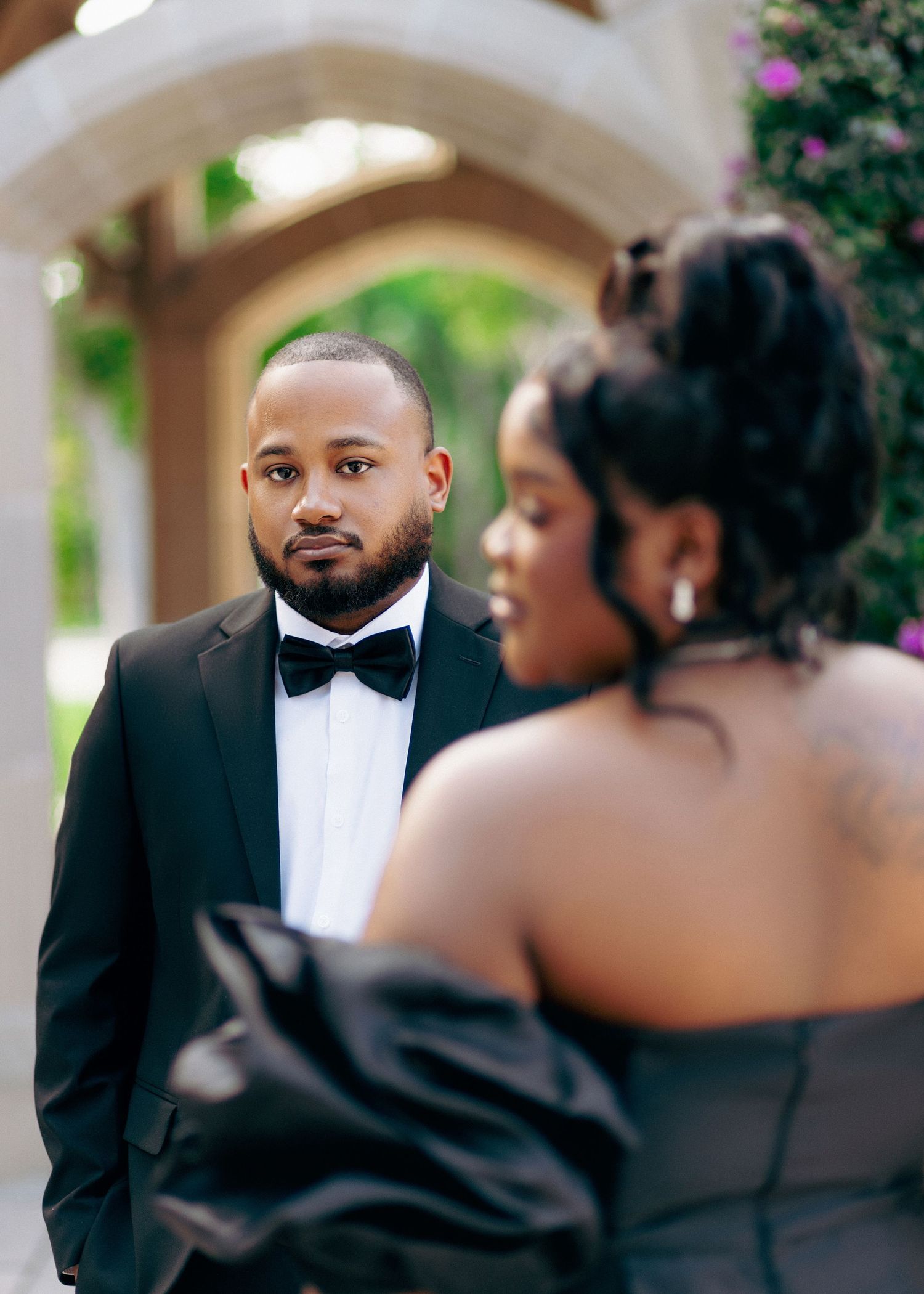 A groom in a black tuxedo gazes at his bride in a strapless black gown during an elegant outdoor engagement session.