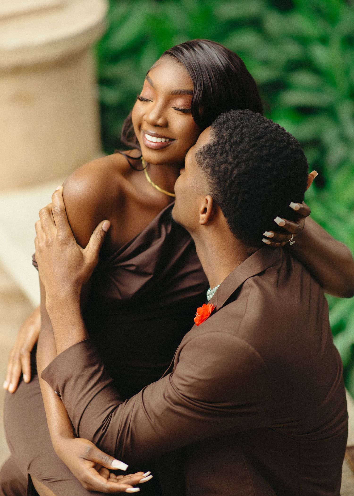 A couple wearing brown outfits share intimate moments in a romantic outdoor photoshoot with green foliage background.