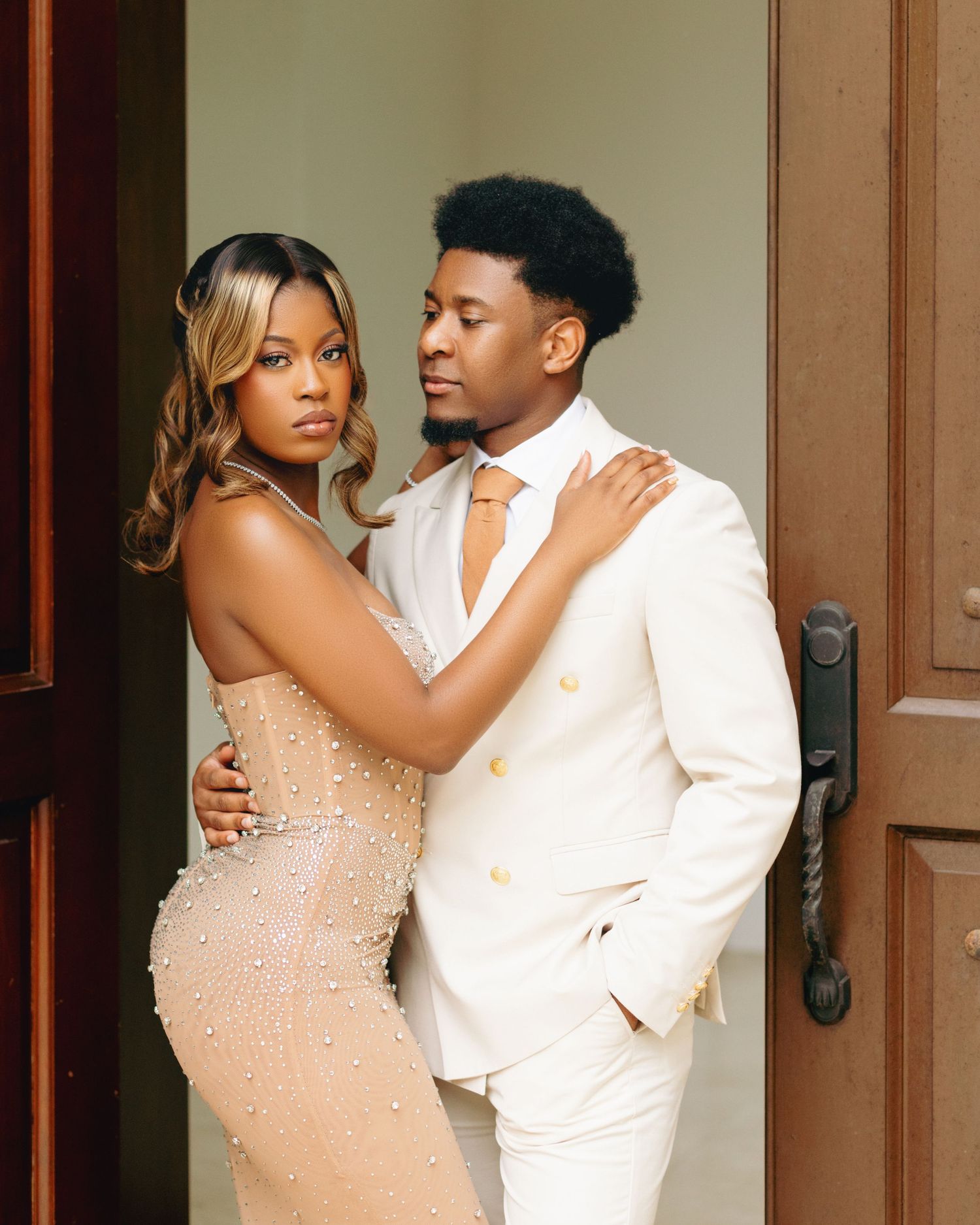 A couple in formal attire share an intimate moment against a wooden door in a romantic photoshoot.
