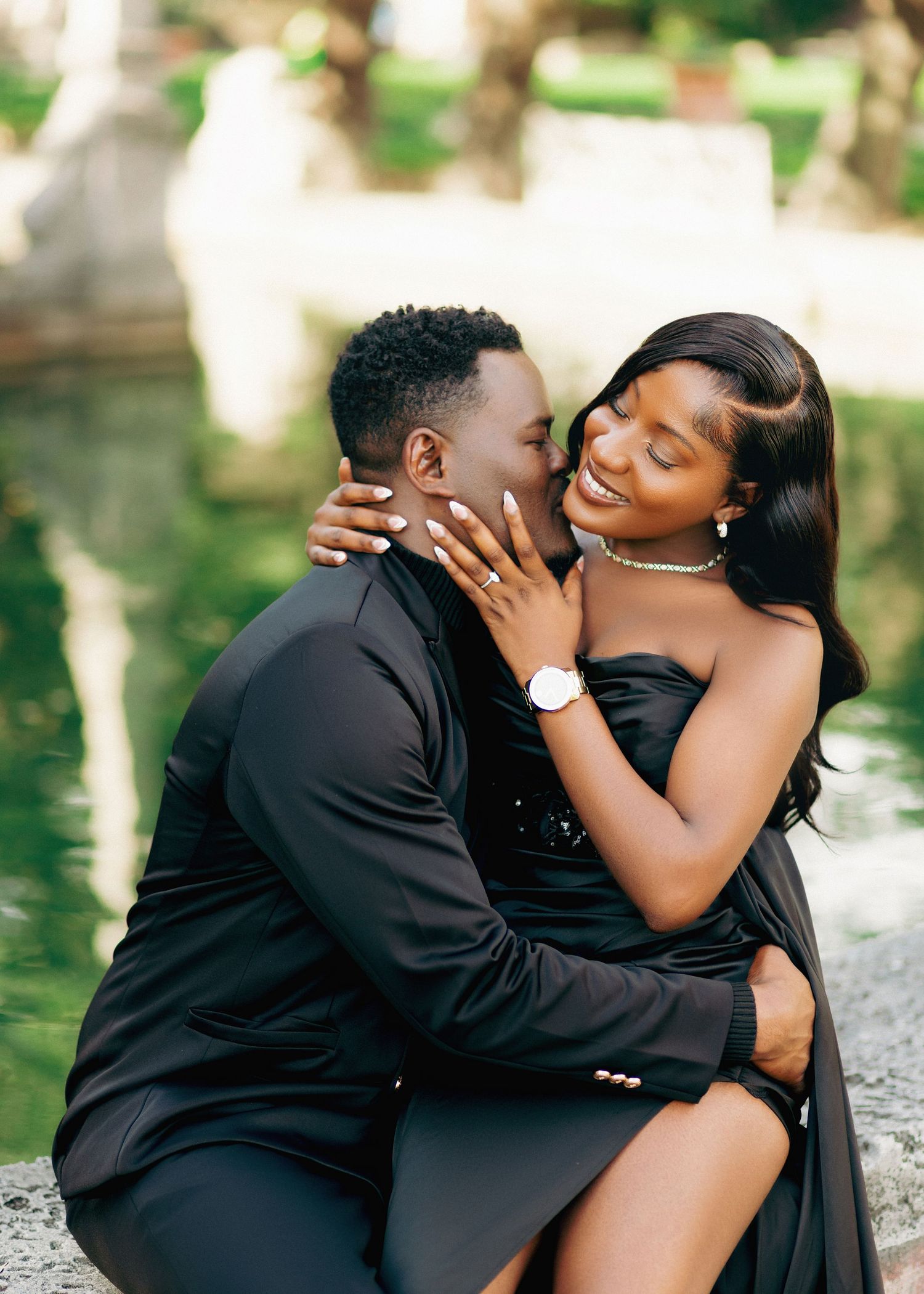 A romantic couple in elegant black attire share an intimate embrace by a reflective pond in an outdoor setting.