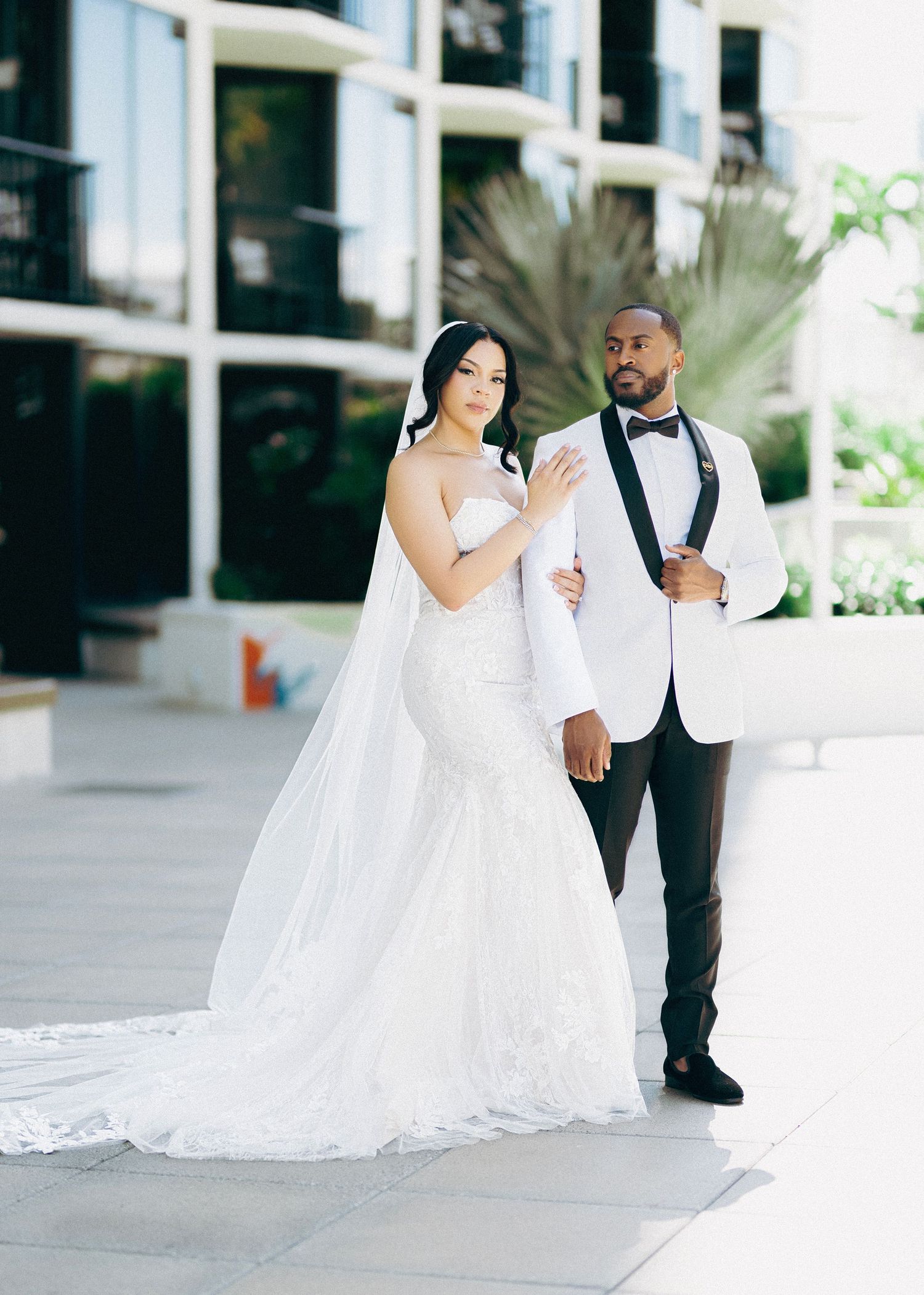 Bride in mermaid wedding gown with veil and groom in white tuxedo jacket pose together outdoors.