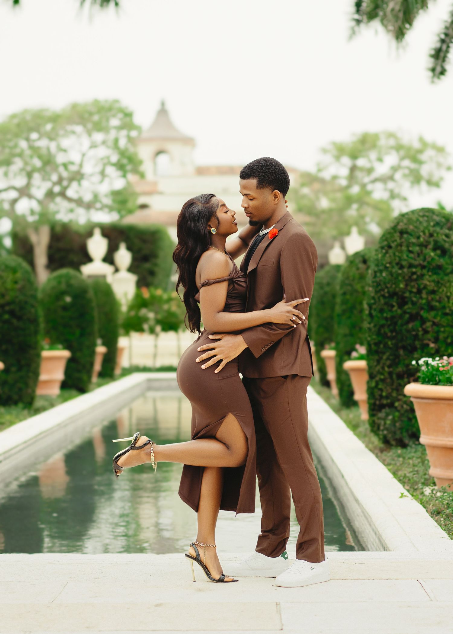 A romantic couple embraces by a reflecting pool in a manicured garden with a church steeple visible in the background.