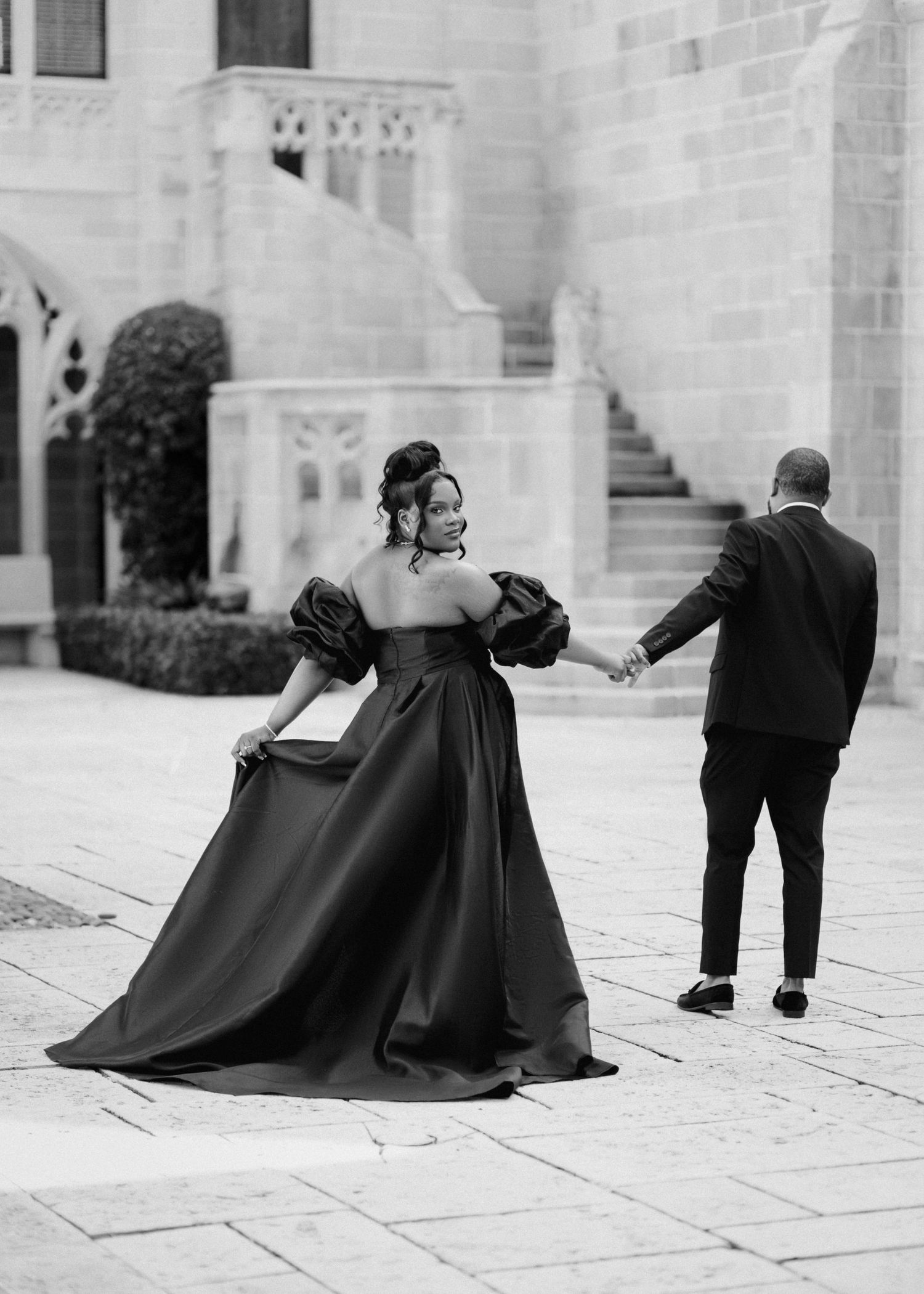 A couple in formal attire poses dramatically on stone steps, with the person in a flowing black ballgown holding its skirt.