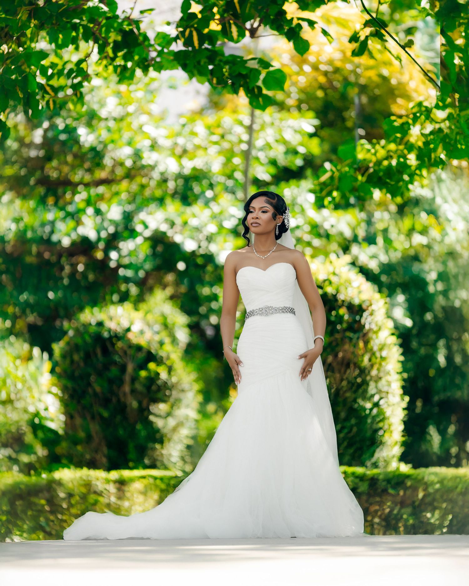 A bride in a strapless white wedding gown with crystal belt poses outdoors under lush green trees in natural sunlight.